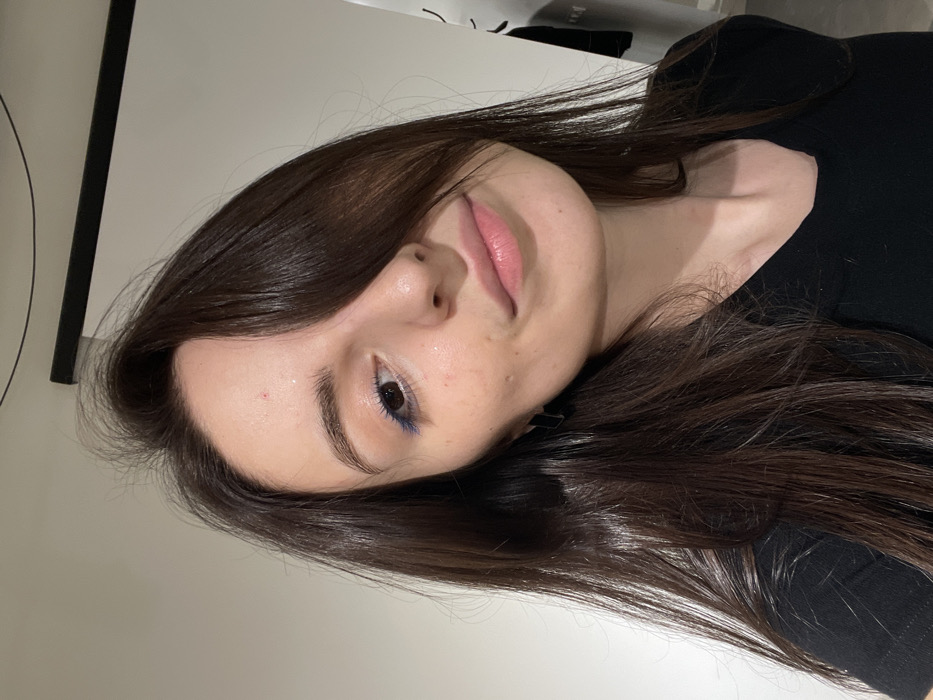 A young woman with light skin and dark brown hair looks at the camera. She wears a black shirt and has blue eyeliner on her lower eyelid. Her lips are a natural pink. The background consists of a white wall and a black rectangular frame. The image is rotated, tilting the subject's head towards the left.