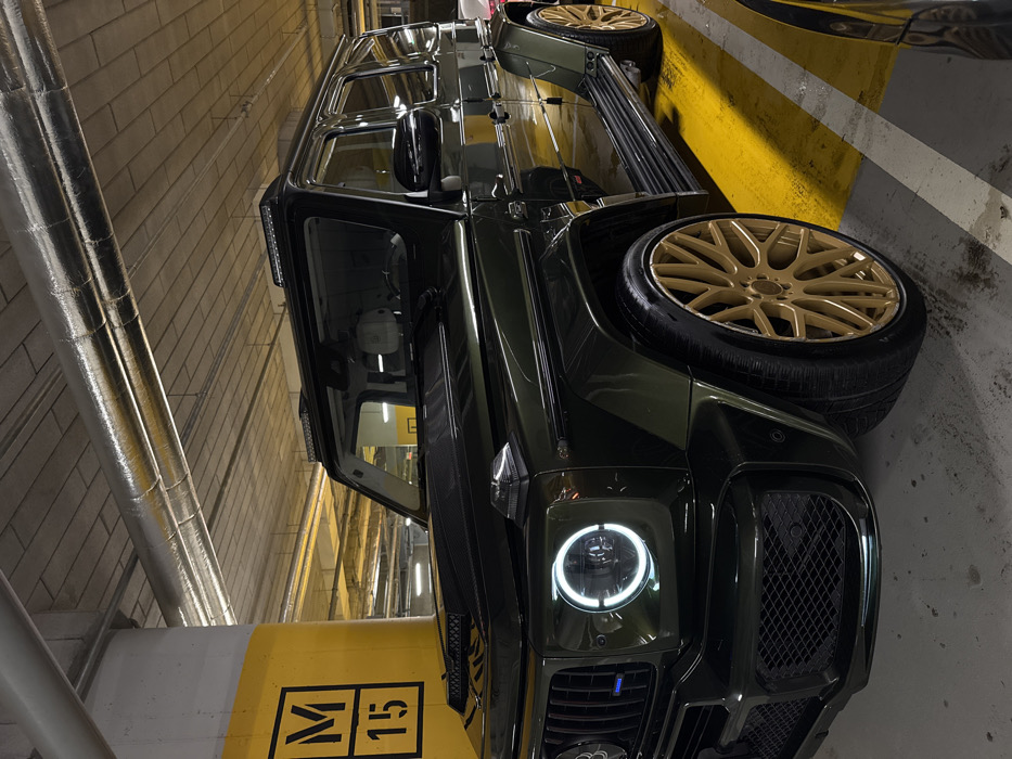 A dark green Mercedes-Benz G-Wagen, possibly an AMG variant, is centrally positioned and parked in an underground garage. The car features prominent gold or bronze multi-spoke alloy wheels with tires. Its circular LED daytime running lights are illuminated, and it has a large, dark mesh grille on the front. Above the windshield, there appear to be integrated LED light bars. The car's deep green paint reflects the artificial lighting of the garage.
The setting is an underground parking facility, characterized by concrete walls with a light brown/beige brick or block pattern in the background. Overhead or along the upper wall, silver insulated pipes are visible. A yellow and white painted concrete pillar stands in the foreground, partially obscuring the car's front. On this pillar, the black alphanumeric text "M 15" is clearly visible, likely indicating a parking bay number. The concrete floor of the garage also features yellow painted lines. No people or specific activity beyond parking are depicted. Given the provided context, the location is Warsaw, Poland.