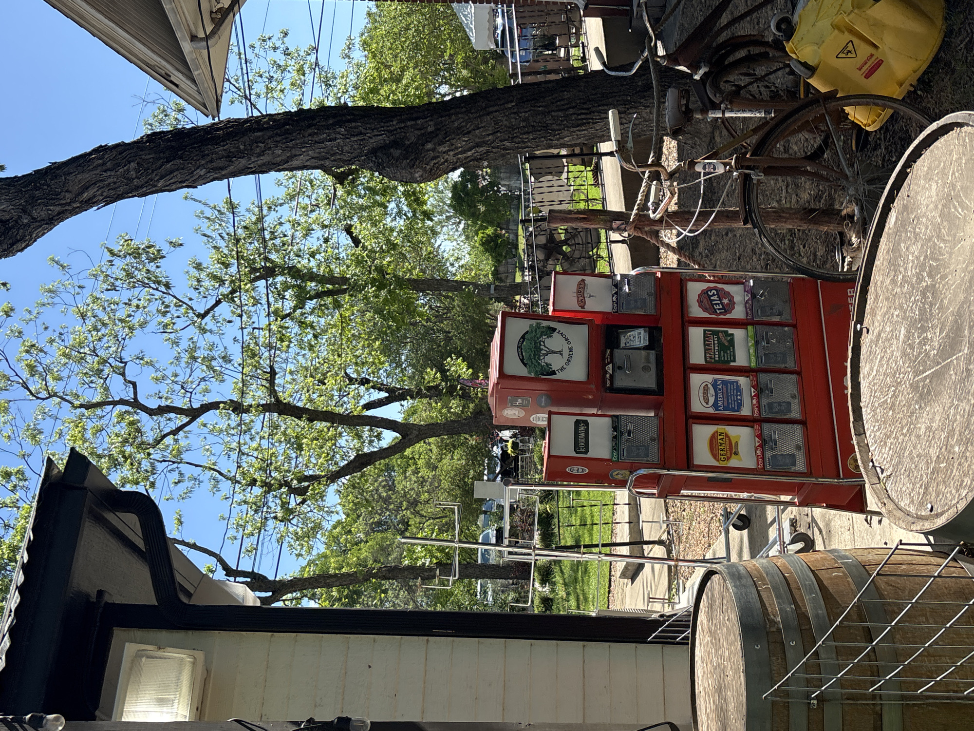 The image depicts a collection of vintage-themed objects in an outdoor setting. The focal point is a red machine with multiple slots, each bearing a different label like "THE GRUENE GROVE", "WURST", "TEJAS", "GERMAN", "AMERICAN", and "ITALIAN BRATWURST", suggesting a theme related to food or culinary culture. A bicycle is leaning against it, with a yellow container attached. A wooden barrel is placed beside the machine.
The scene is outdoors, evidenced by trees and a patch of green grass. Part of a building is visible on the left side. The items and surrounding environment appear to have a vintage or rustic style.
Given the presence of "THE GRUENE GROVE" and "TEJAS", it is reasonable to infer the location as Gruene Historic District in New Braunfels, Texas, which is known for its historic buildings and landmarks.