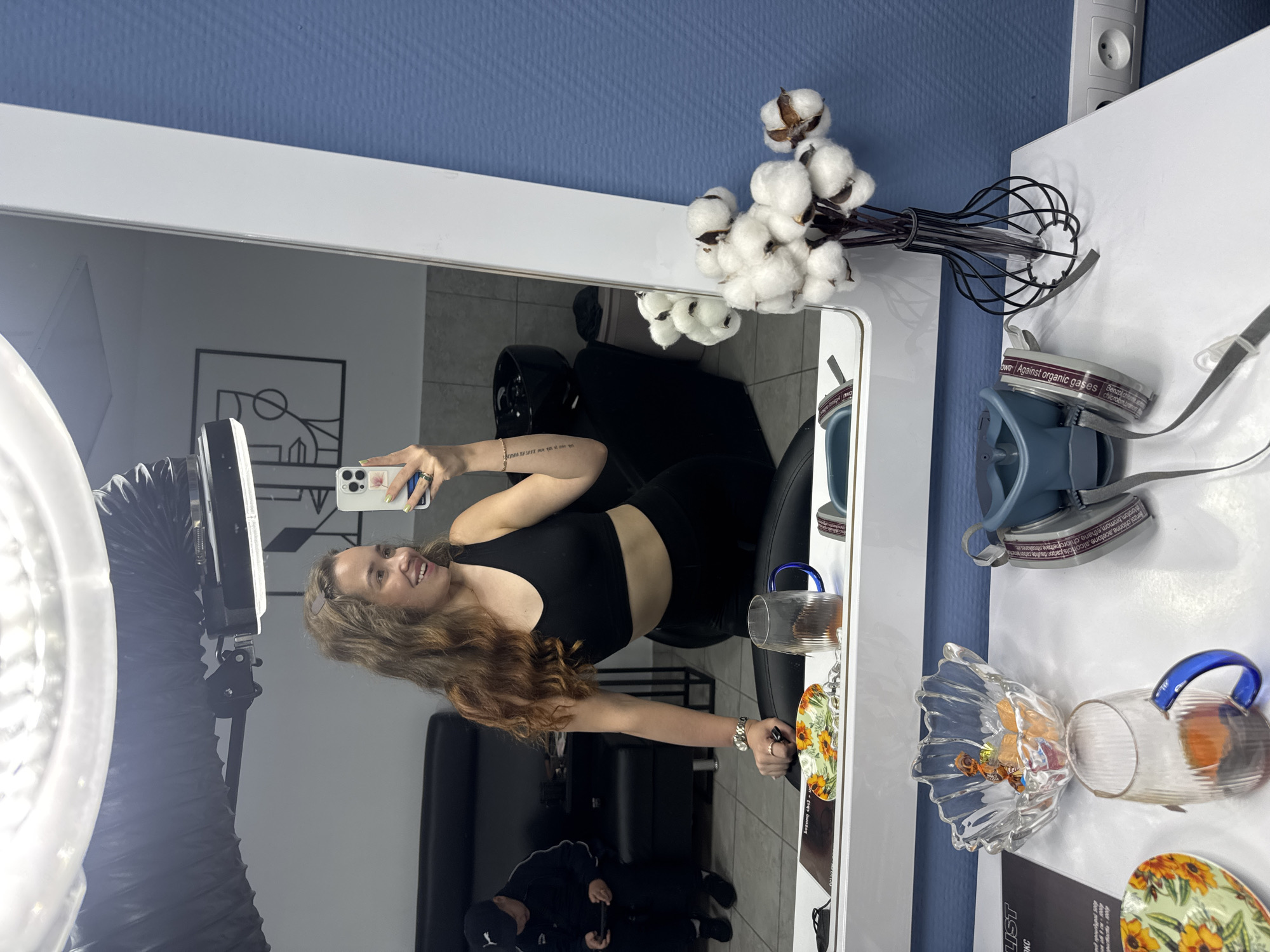 A young woman is captured taking a mirror selfie in what appears to be an indoor setting, possibly a salon or studio, in Chaikovsky, Russia. She stands smiling, holding a smartphone with a floral-patterned case and light-colored nails, reflecting in a large, white-framed mirror. She has long, wavy brown hair, wears a black crop top, and has a tattoo visible on her right forearm.
Reflected in the mirror behind her, a man is partially visible, seated on a black couch or chair, also seemingly looking at a phone. A large ring light with a flexible tube arm is prominently visible to the left, illuminating the scene.
On a white surface beside the mirror, several objects are arranged. A bouquet of cotton plants in a black wire vase adds a decorative touch. Nearby, a protective mask or respirator is placed, featuring text such as "room, broom.ethane.choгontroalkanes," "Benzoichionine, acetone, alcohols parodie," "Benzol chlorme," and "own Against organic gases chlorambrano," indicating filter specifications or warnings. Further along the surface are various clear glass items, including cups or mugs (one with a blue handle), a small fluted glass bowl containing what appears to be candy, and decorative plates or coasters with a sunflower motif. A white electrical outlet is visible on the blue wall above. The overall impression is a modern, well-lit space.