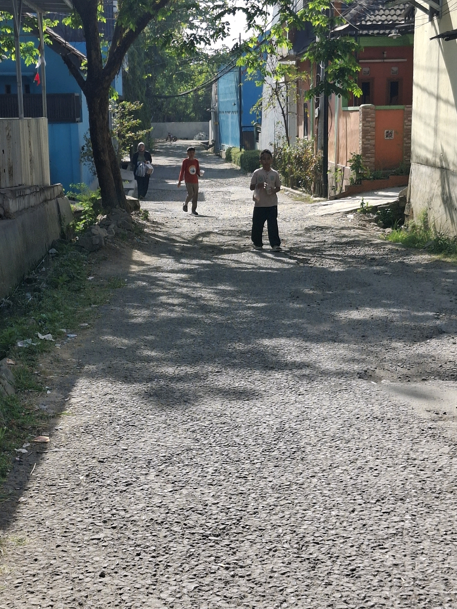 This image captures a street scene in Karangtengah, Indonesia, under bright sunlight, casting strong shadows across a textured, gravel-covered road. On the right side of the road, a person stands facing the viewer, wearing dark pants and a light-colored short-sleeved shirt, holding a clear plastic bottle in their right hand. Further up the street, a child in a red long-sleeved shirt and dark pants is running towards the camera, holding a white cup. To the left of the running child, a woman wearing a hijab and dark clothing is walking away from the camera, carrying a striped bag and a white box. The street is flanked by buildings: on the left, a blue building with a light-colored wall and a metal fence is visible, and on the right, several residential buildings with orange and beige facades and dark tiled roofs are present. Vegetation, including trees and bushes, grows alongside the buildings and in patches on the side of the road, with some litter visible on the grassy verge to the left. The overall impression is a quiet, everyday moment in a residential urban area.