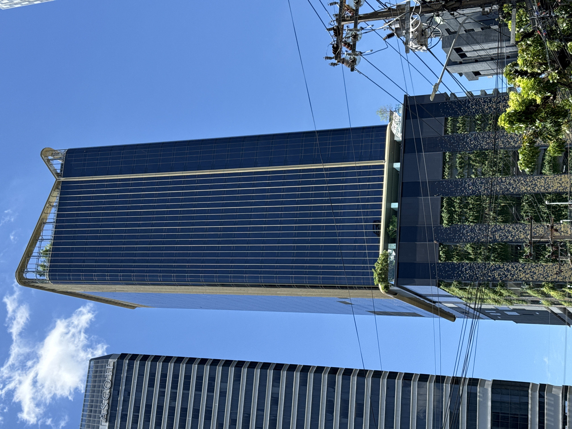 The image shows a modern skyscraper in Bangkok, Thailand, against a clear blue sky with a few scattered clouds. The skyscraper is tall and slender, with a dark glass facade and gold accents. The building features a green vertical garden on its lower levels and a rooftop garden. To the left of the main skyscraper, another taller building with horizontal stripes is visible, displaying the "ASCOTT" logo at the top. Overhead power lines and a utility pole with electrical equipment are present in the foreground, with lush green trees also visible, suggesting an urban environment. The overall impression is one of contemporary urban architecture and infrastructure.