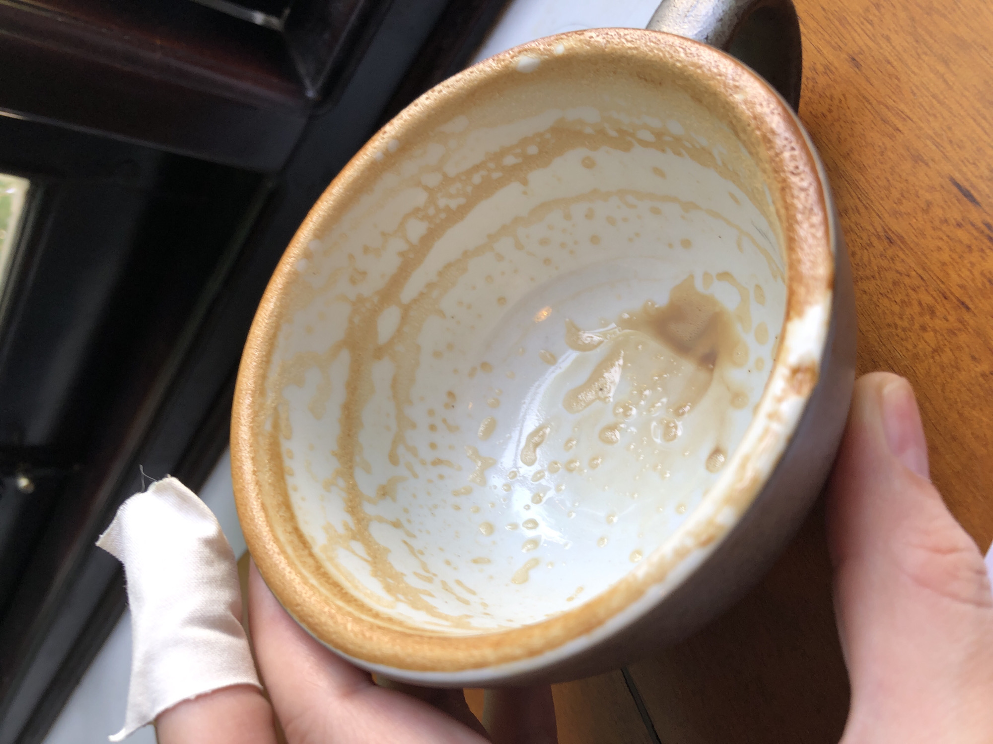 A close-up, top-down shot shows the interior of a coffee cup with remnants of coffee. The cup is white with a brown rim, and streaks of brown coffee are visible on the inside walls, indicating it has been used. A person's thumb and forefinger are holding the cup from the left side, and a small piece of white fabric is seen on the index finger. The cup rests on a wooden surface, possibly a table. In the background, a dark wooden structure with a window is visible. The image does not provide specific details about the location in Da Nang, Vietnam, other than it appears to be an indoor setting with natural light coming from a window.