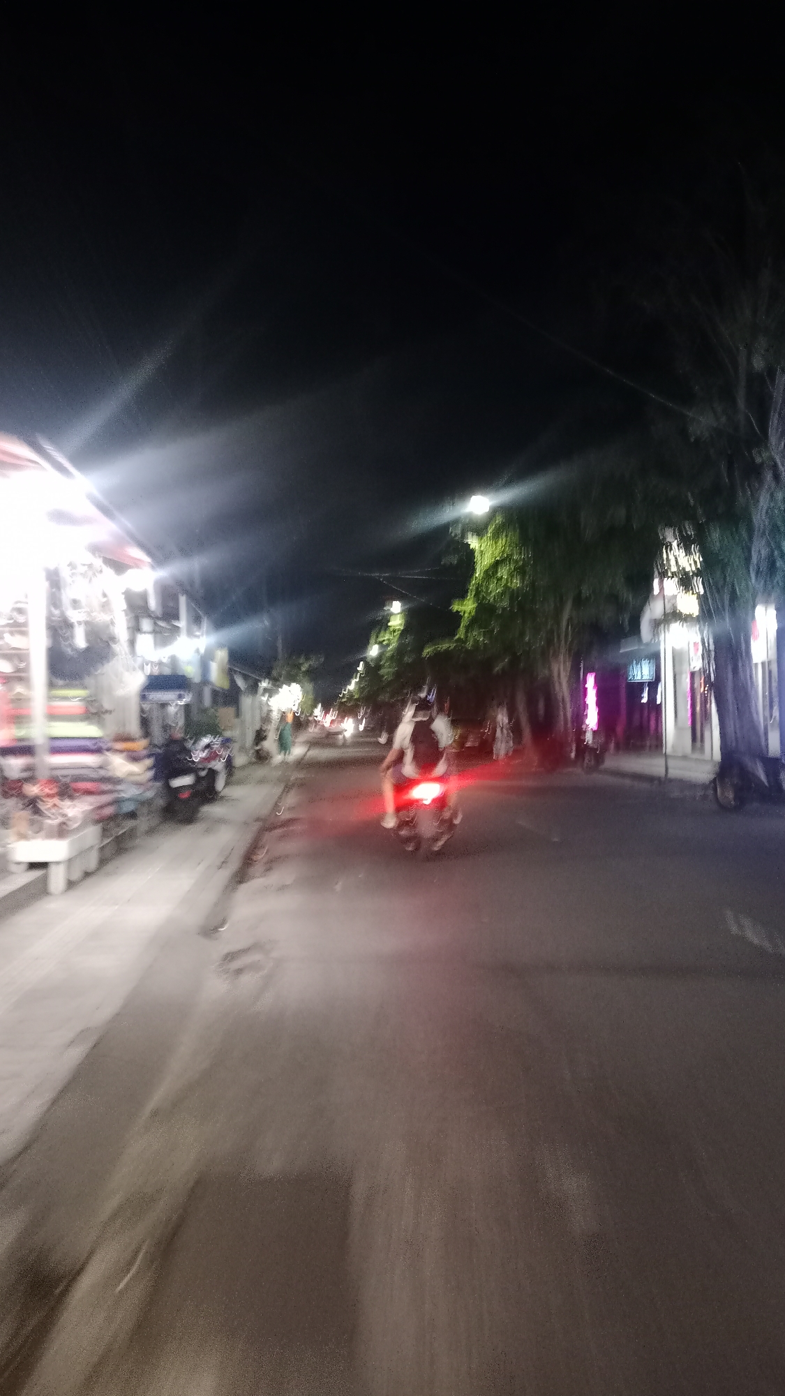 The image shows a street scene at night in Kuta, Indonesia. A person is riding a motorcycle on the right side of the road, with their red taillight illuminated. The motorcycle and rider are slightly blurred, suggesting motion. On the left side of the road, there are shops or stalls with items displayed, illuminated by bright lights. Trees line the right side of the street, and streetlights are visible along the road, casting light on the pavement and surroundings. The overall impression is that of a bustling street at night.