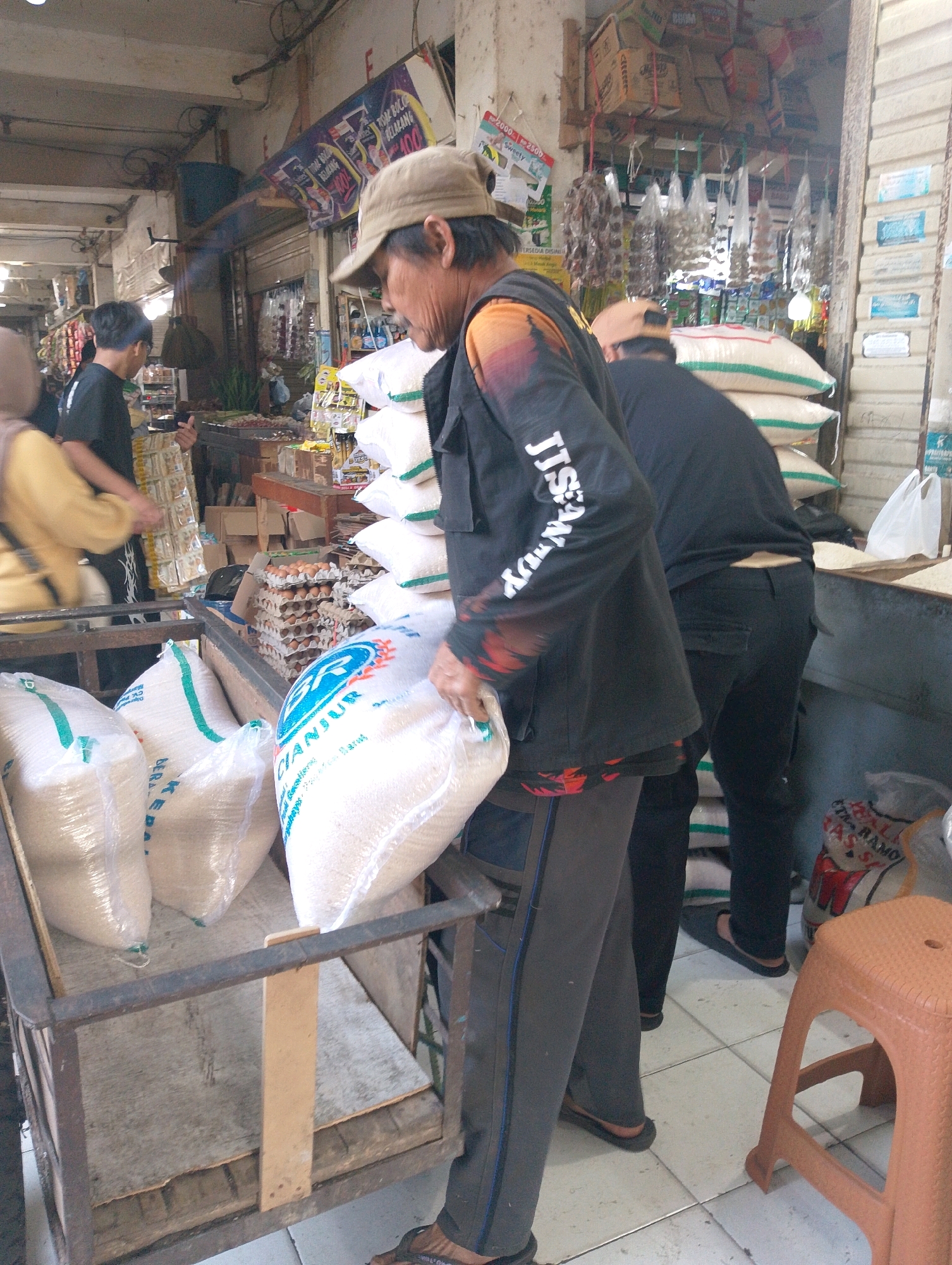The image depicts a market scene in Lembang, Indonesia, with a focus on a stall selling sacks of rice and other goods. In the foreground, a man wearing a dark jacket with "JJSEANIM" written on the sleeve and a beige cap is in the process of lifting a large, translucent sack filled with white granular material, presumably rice. He is wearing dark trousers and flip-flops. To his right, another person, partially visible and wearing a black t-shirt and black trousers, is also attending to the sacks. Behind them, a young man in a black t-shirt and glasses is seen arranging items on a shelf. The stall displays multiple stacks of white sacks, some with green trim, and a cart loaded with more sacks. In the background, shelves are filled with various packaged goods, including boxes and what appear to be dried food items hanging from the ceiling. The environment is that of a bustling market with a tiled floor and overhead structures suggestive of a covered market area. A small, orange plastic stool is visible to the right. The overall impression is a typical retail setting for everyday commodities.