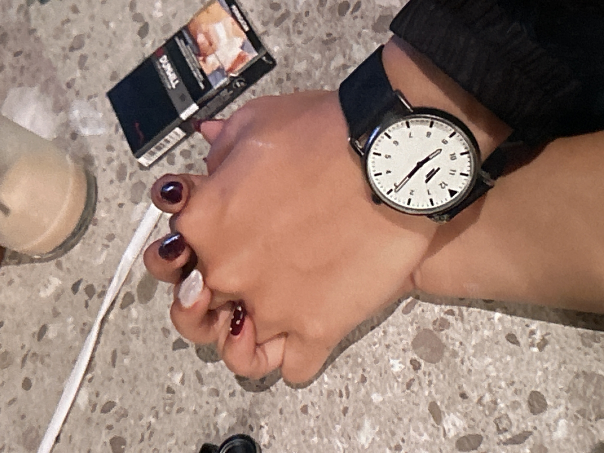 The image depicts a close-up, overhead view of two pairs of hands interlocked. One hand, adorned with dark red nail polish and a white accent nail, is holding the other hand. The second hand, wearing a black watch with a white face and black band, is also visible. Beside the hands, a pack of Dunhill cigarettes is placed on a speckled, gray countertop. A white straw and a portion of a beverage in a glass are also present in the background, suggesting a casual setting. The scene is likely in Depok, Indonesia.