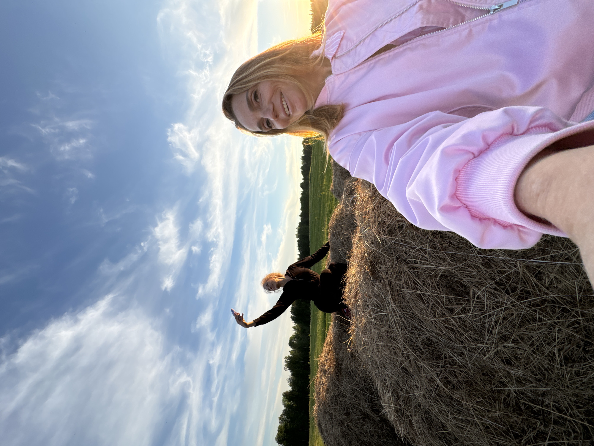 The image captures an outdoor scene in a rural setting, illuminated by the warm, golden light of late afternoon or early evening. The sky overhead is a bright blue with scattered white, wispy clouds, indicating clear weather.
In the foreground, a woman with long, light brown hair, wearing a vibrant pink jacket, is taking a selfie. She is smiling broadly towards the camera, her face and hair beautifully lit by the low sun. Her arm is visible on the right, extending to hold the camera.
Further back, positioned on a large, textured hay bale, another woman with lighter hair, dressed in dark clothing, is seen posing. She holds a smartphone in her outstretched hand, seemingly capturing a photo or video of the landscape or herself. The hay bales are prominent, made of dried straw. Beyond them, a green field extends towards a distant treeline. The overall mood is cheerful and relaxed, depicting an enjoyable outdoor moment in the region of Стеклянный, Russia. No visible text is present in the image.