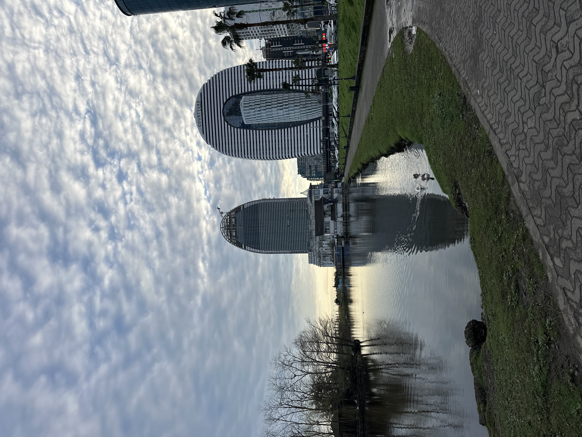 The image shows a waterfront scene in Batumi, Georgia. Notable buildings, including the Courtyard by Marriott, are reflected in the water. A paved path runs along the edge of the water, with grass and trees lining the banks. Two ducks swim in the water. The sky is filled with clouds.