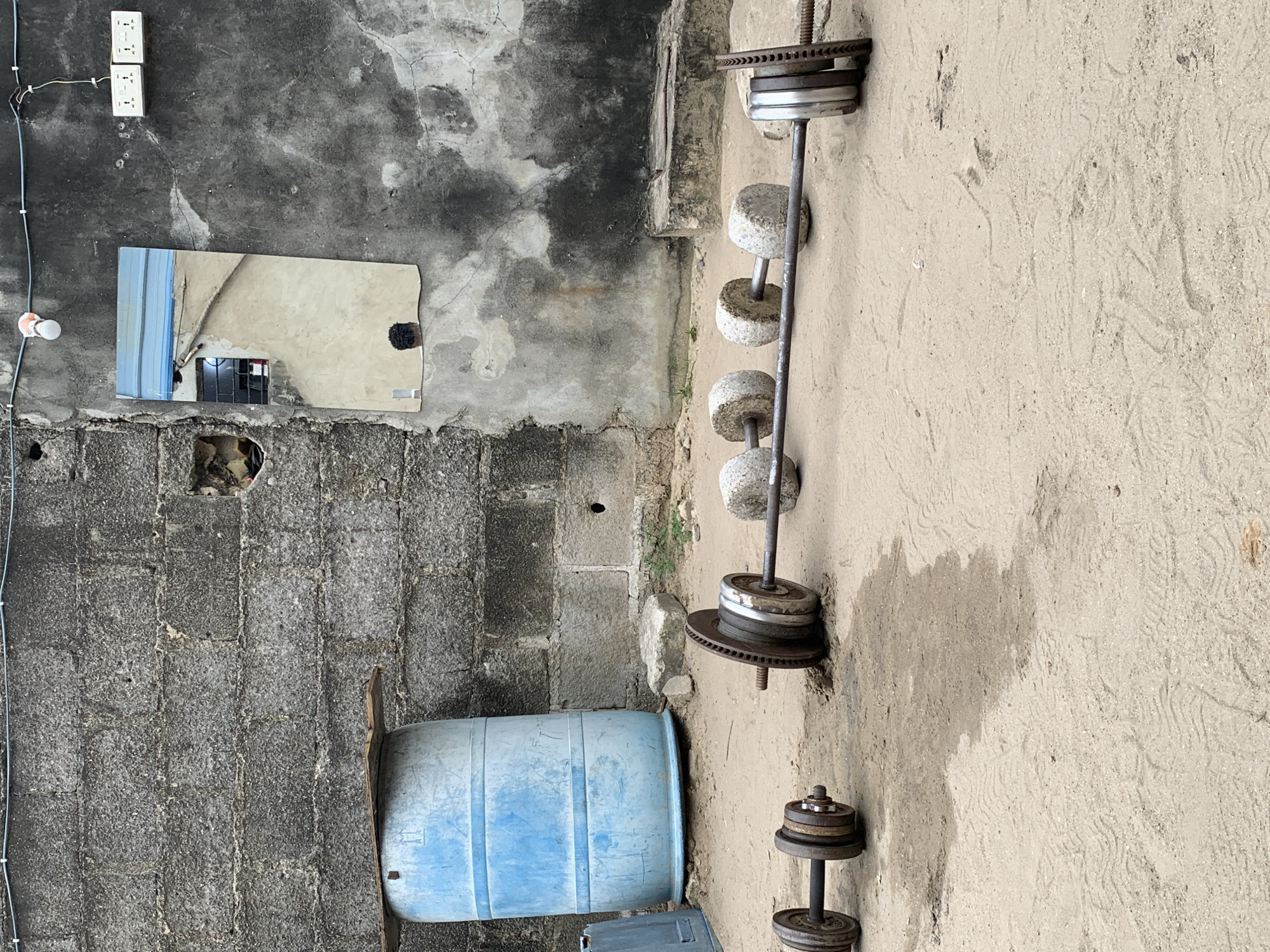 An outdoor courtyard in Ado, Nigeria, features a grey, rough-textured concrete block wall forming the background. A large, weathered blue plastic barrel stands to the left, with a piece of wood resting on top. To the right of the barrel, a mirror is affixed to the wall, reflecting a pale blue structure and a window with bars. Two electrical outlets are visible on the wall above the mirror.
In the foreground, the ground is sandy and appears somewhat damp in patches. Two sets of weightlifting equipment are present. On the left, a dumbbell with multiple metal plates and collars rests on the sand. To its right, a barbell lies on the ground, loaded with circular weights that appear to be made of concrete. One end of the barbell has metal plates stacked on it. A large, rough stone is situated near the barbell. The overall scene suggests a makeshift outdoor gym.
