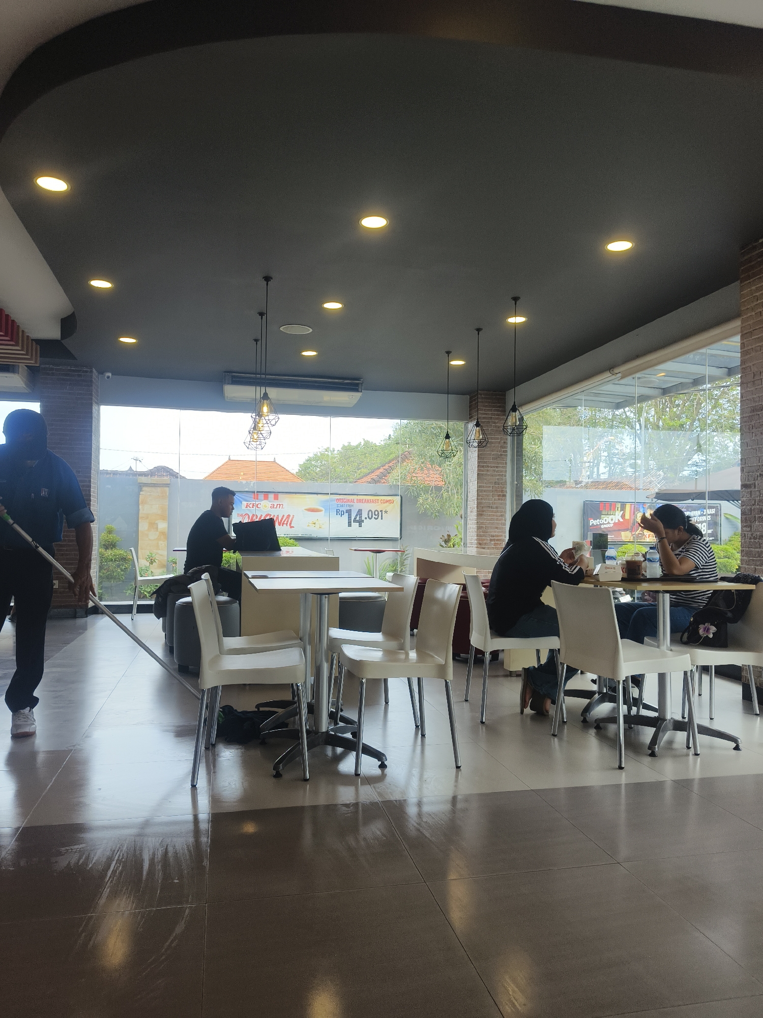 In Denpasar, Indonesia, a man wearing a dark blue shirt and dark pants is mopping a shiny tiled floor inside what appears to be a KFC restaurant. The restaurant has large glass windows offering a view of lush greenery and buildings outside. Several tables with white chairs are set up for dining. In the background, patrons are seated at tables, some of whom are engaged in conversation or using laptops. A large advertisement for KFC is visible on a screen or poster behind the patrons. The ceiling is dark gray with circular embedded lights, and there are hanging pendant lights above some of the tables. The overall scene suggests a casual dining environment during daylight hours.