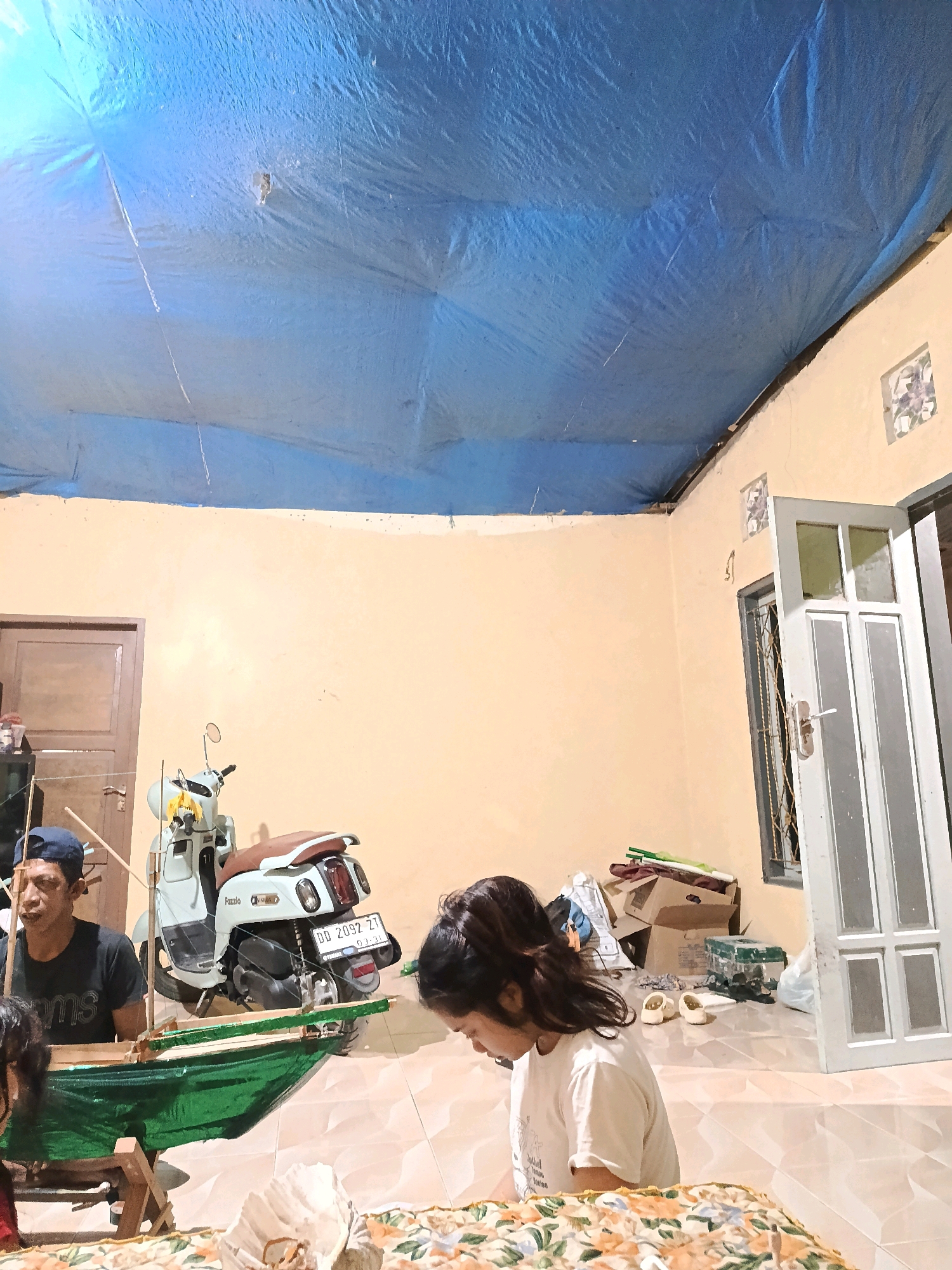 The image depicts an indoor scene, possibly a living area or workshop, in Bulukumba, Indonesia. A blue tarp is stretched across the ceiling, creating a makeshift roof. The walls are painted a pale yellow. In the foreground, two individuals are partially visible. One is an adult male wearing a dark t-shirt and a blue baseball cap, seated and looking down at a green-and-brown model boat. He appears to be working on it. To his right, a young girl with dark, long hair is also looking down, seemingly at something on the floor. She is wearing a white t-shirt.
A light-colored scooter with a brown seat is parked in the middle ground. The scooter has a license plate that reads "DD 2092 ZT". Behind the scooter, there are several cardboard boxes and other miscellaneous items. To the left, a closed wooden door is visible. To the right, a white door with glass panes is ajar, revealing a window with a wire mesh. The floor is tiled with a reflective, light-colored pattern. The overall impression is of a functional, perhaps temporary or multi-purpose, domestic space.
