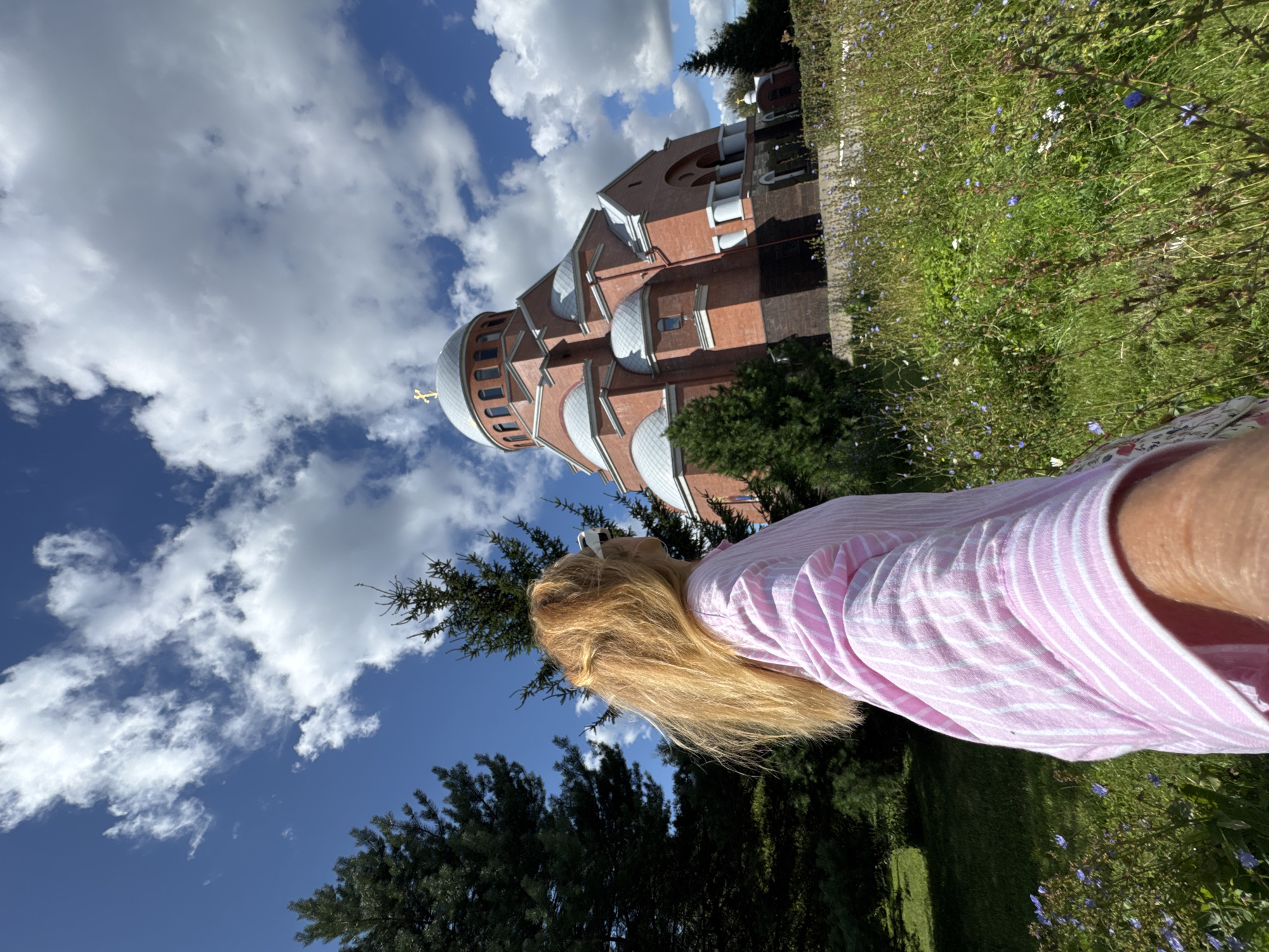 A woman with long, blonde hair is standing in a field of wildflowers, looking towards a large, red brick church with a dome and a golden cross. The church is a prominent feature in the background, with its imposing architecture and several windows visible. The sky is blue with fluffy white clouds. The woman is wearing a pink and white striped shirt. The scene appears to be outdoors on a sunny day, likely in a park or garden area surrounding the church. The overall impression is peaceful and scenic.