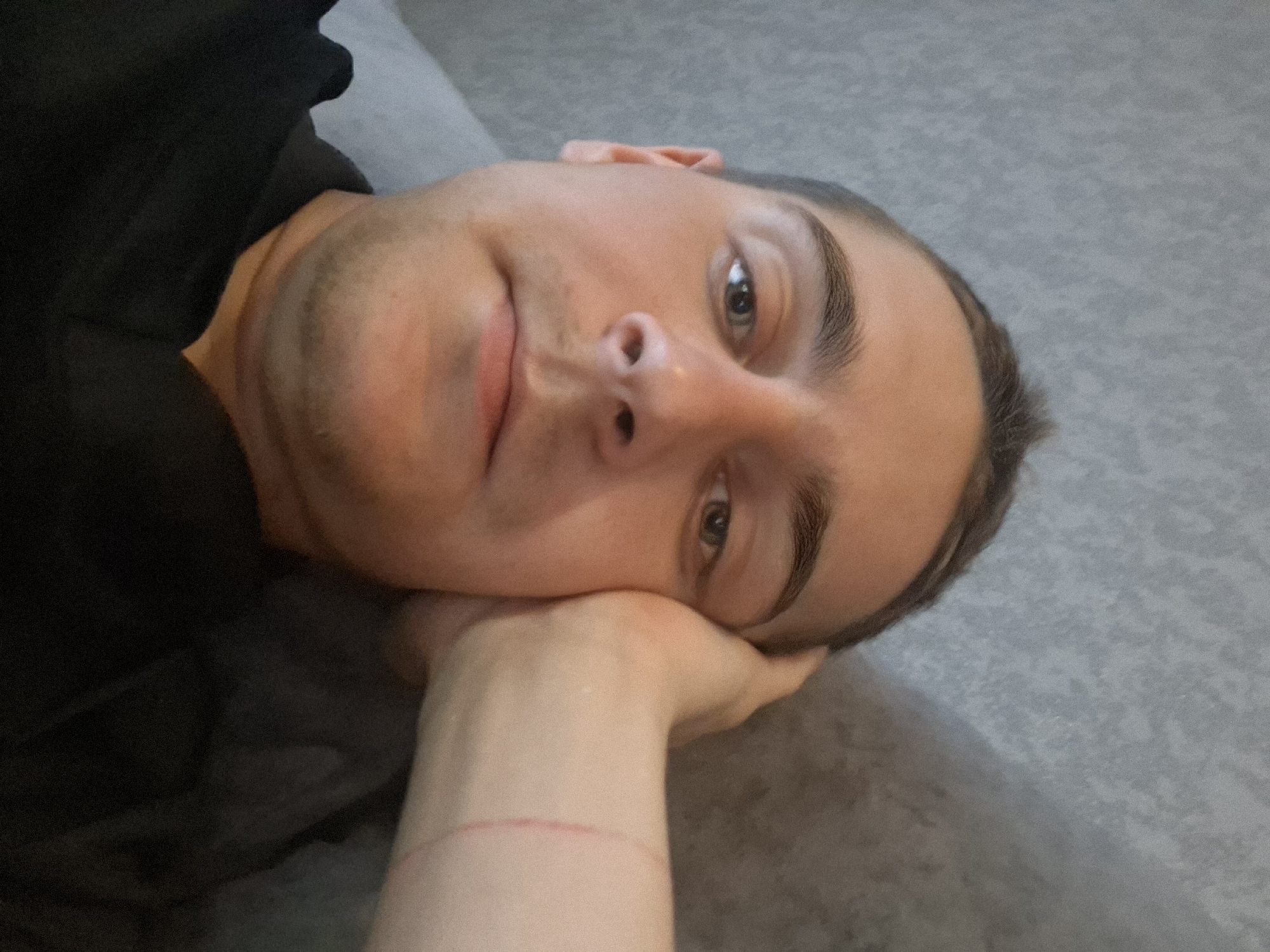 A man is lying on a gray surface, possibly a couch or bed, wearing a black shirt. His head rests on his hand. The location is Unknown, Unknown.