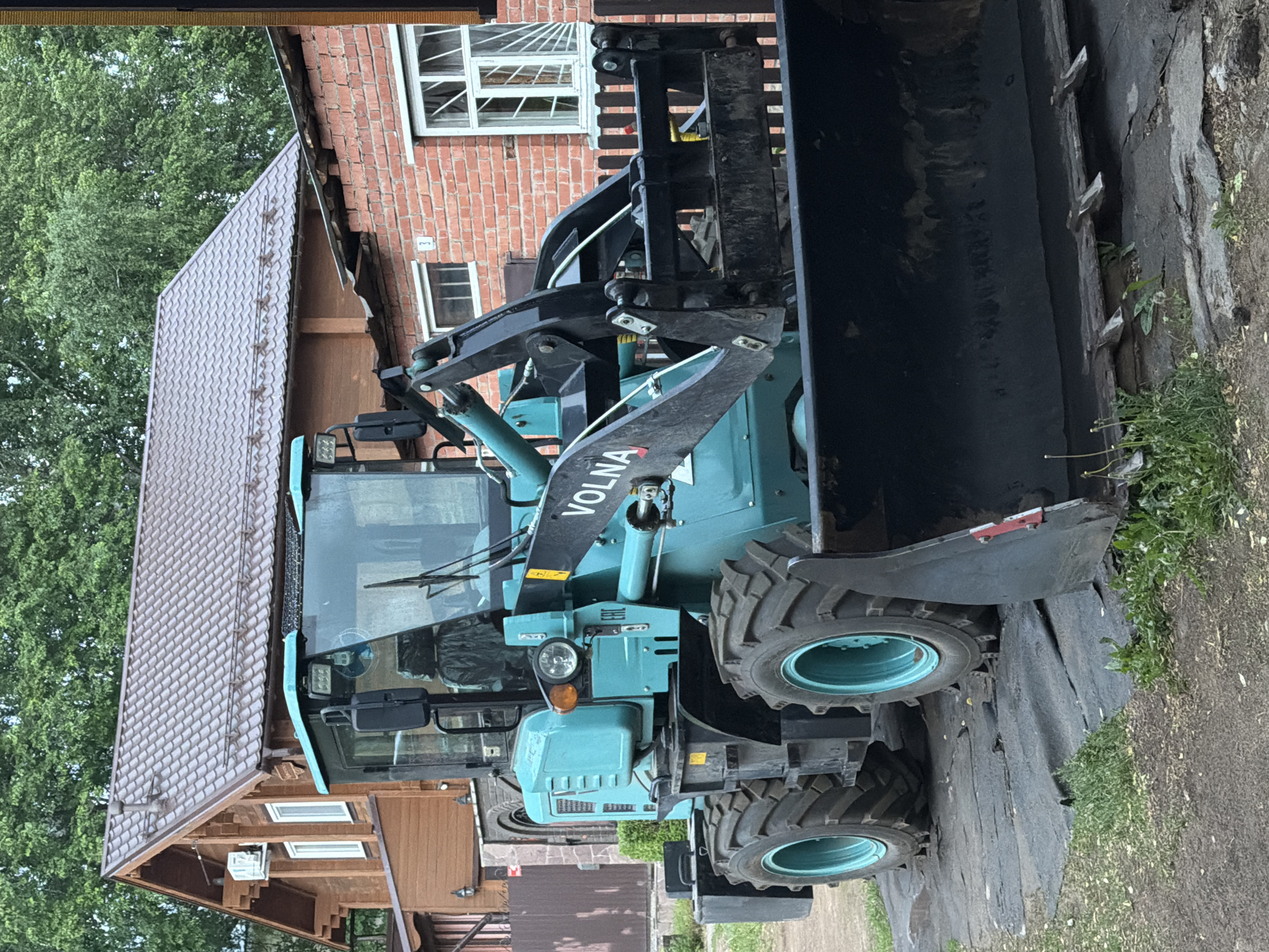 A pale blue VOLNA front loader with a black bucket stands in front of a brown-and-red brick building in Votkinsk, Russia. The building has a brown shingle roof and white-framed windows. The front loader's tires are teal and black, and the bucket is elevated. The ground in front of the loader is covered with a black tarp and some grass. Green trees are visible in the background.