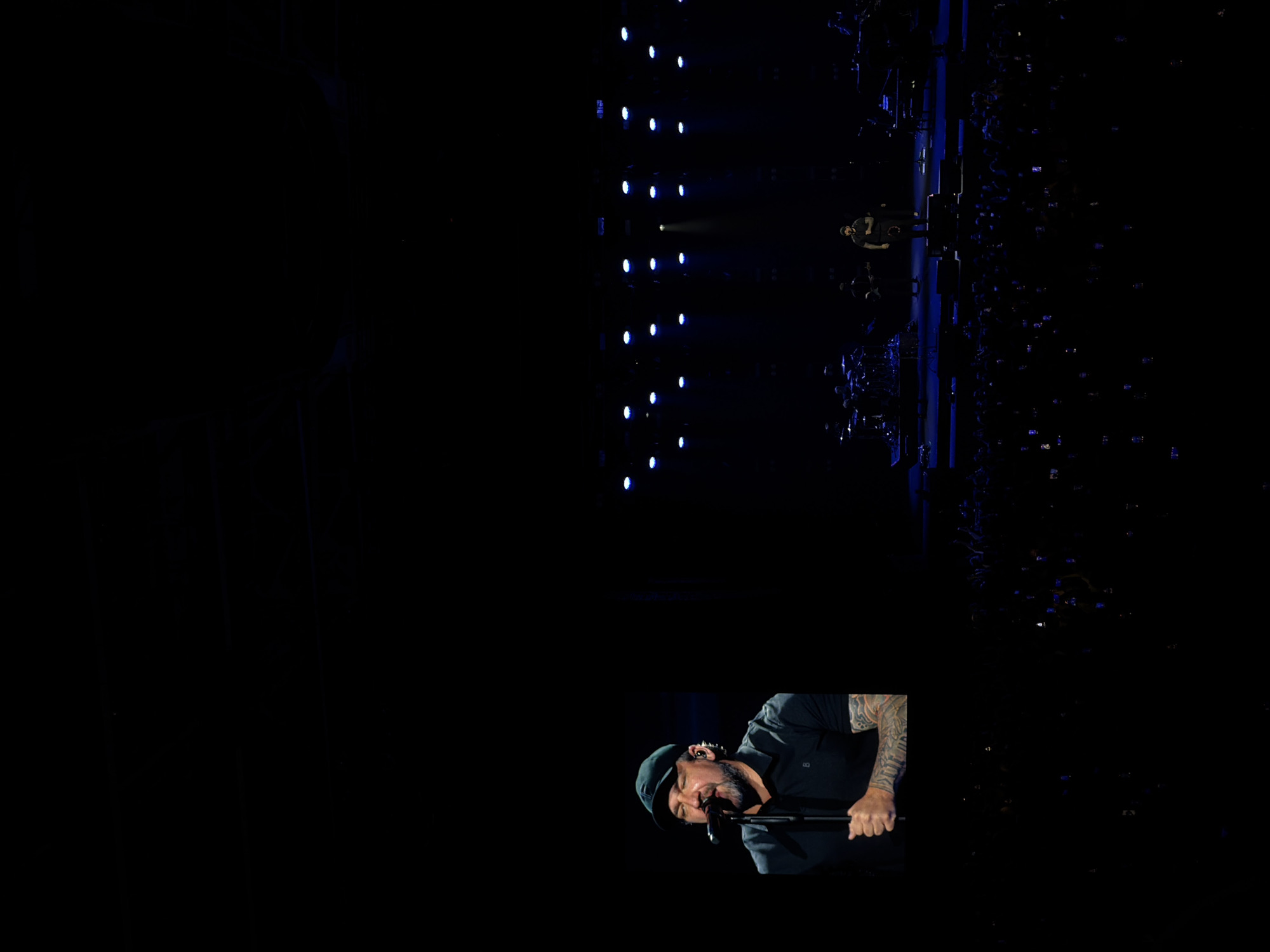 The image captures a musical performance. A male singer is positioned in the foreground, leaning into a microphone. He wears a cap and has tattoos on his exposed arm. In the background, a stage setup with multiple lights is visible, along with a large crowd of people, some of whom are holding up their phones. This likely takes place during a live music event, potentially in a venue within Krasnoyarsk, Russia, though the exact location cannot be determined from the image alone.