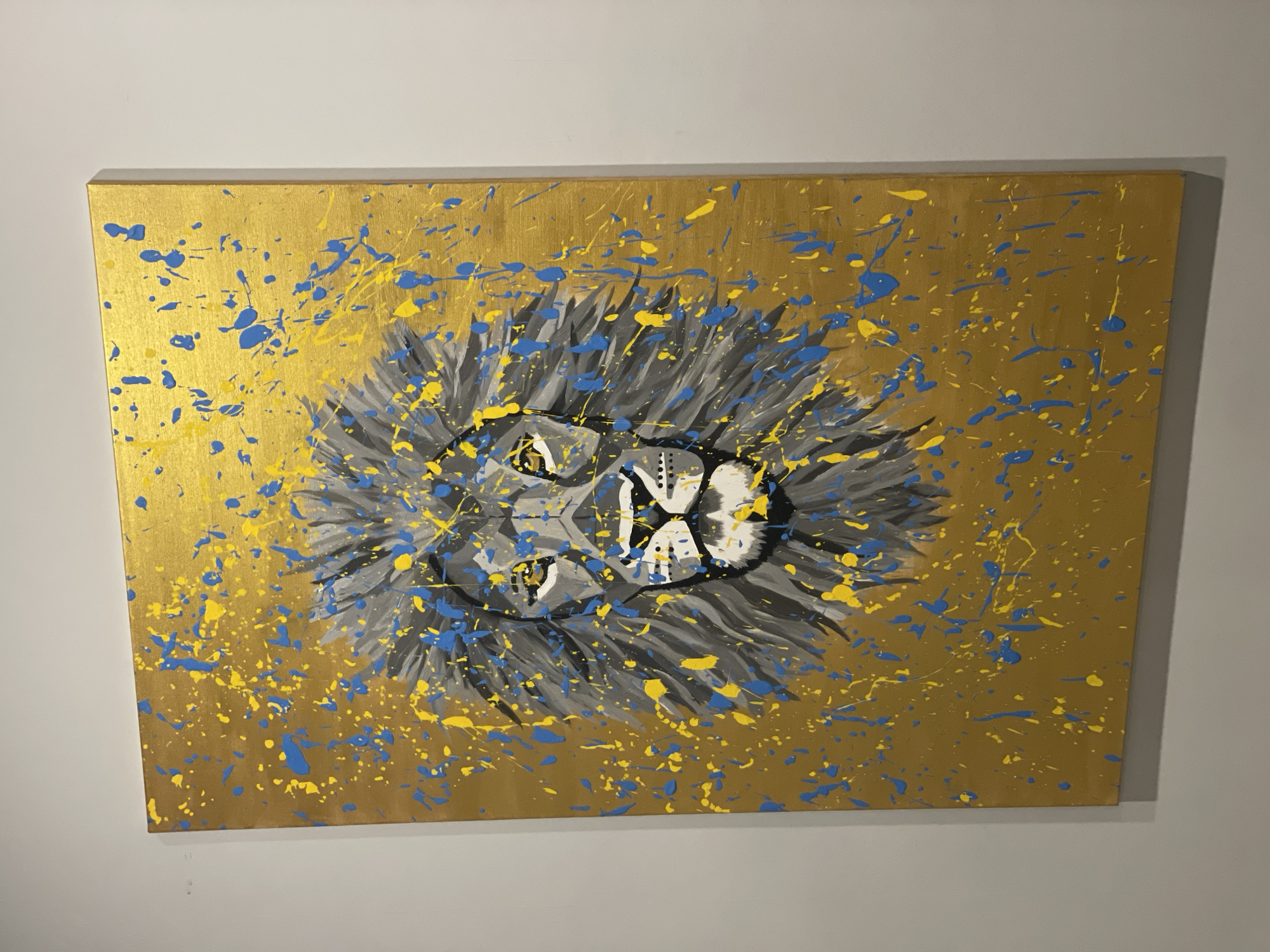 A painting of a lion's face with a gray mane on a gold background with yellow and blue paint splatters hangs on a light gray wall. The painting is rectangular and appears to be a canvas. No information indicates that the painting is in Krasnoyarsk, Russia.
