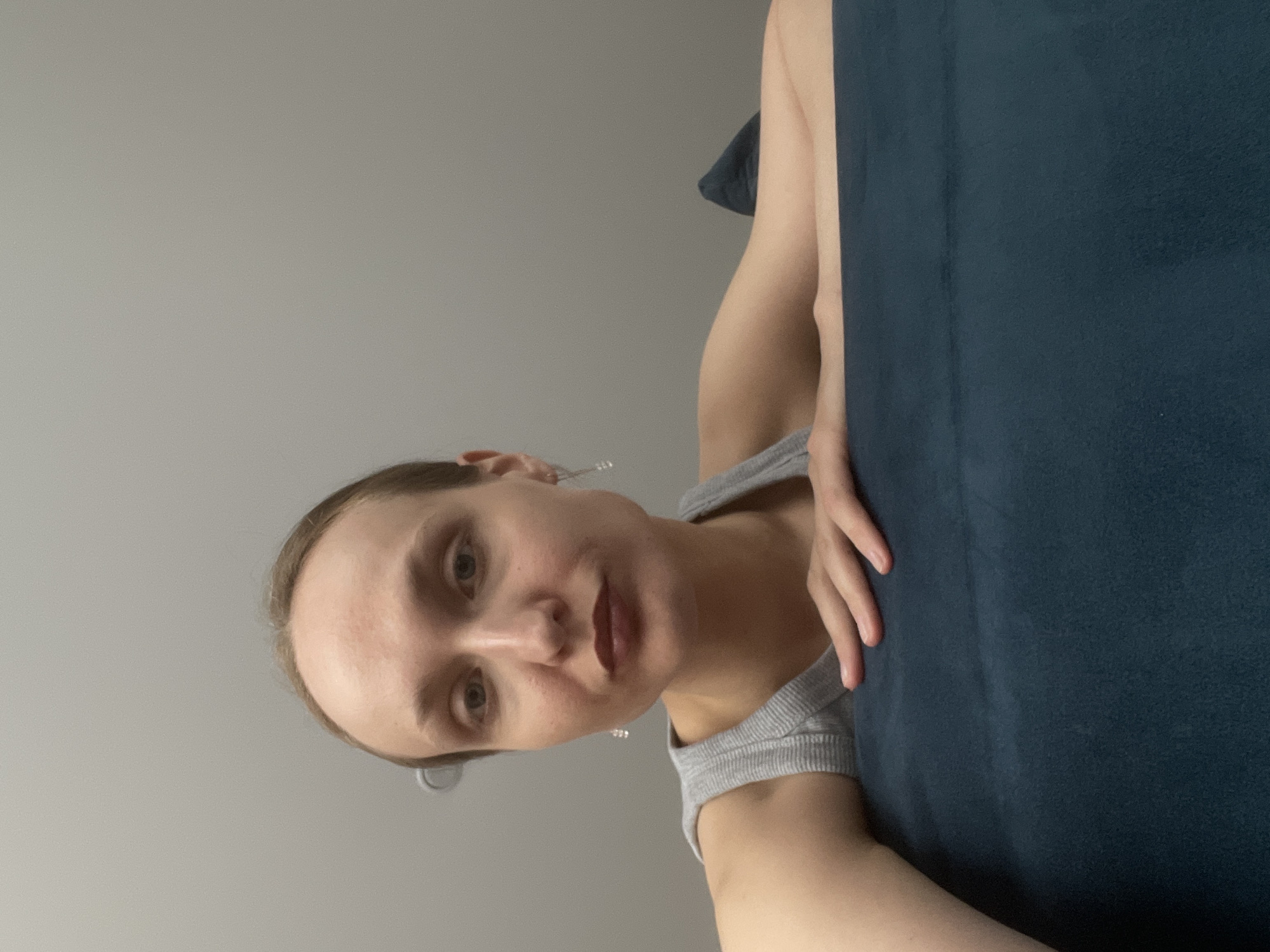 A woman with fair skin and light brown hair is visible from the chest up, wearing a gray ribbed tank top. She is holding a large, dark blue pillow. The background is a light gray wall. No elements indicate the location of Sepych, Russia.