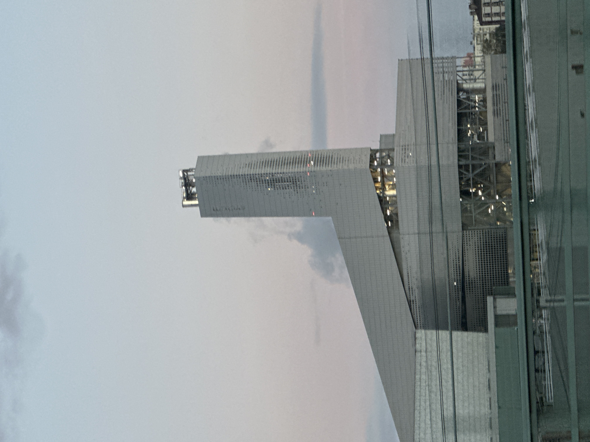 The image shows a modern building, likely an office tower or mixed-use development, viewed through a window. The building's design features a cantilevered section, giving it a distinctive and somewhat futuristic appearance. The exterior material appears to be a light-colored metal or concrete, with a pattern of perforations.
The sky is overcast, creating a muted light. Other buildings are visible in the background, suggesting an urban environment. The window through which the image is taken adds a layer of reflection and distortion.
The building is the French Ministry of Armed Forces, located in the 15th arrondissement of Paris, France.