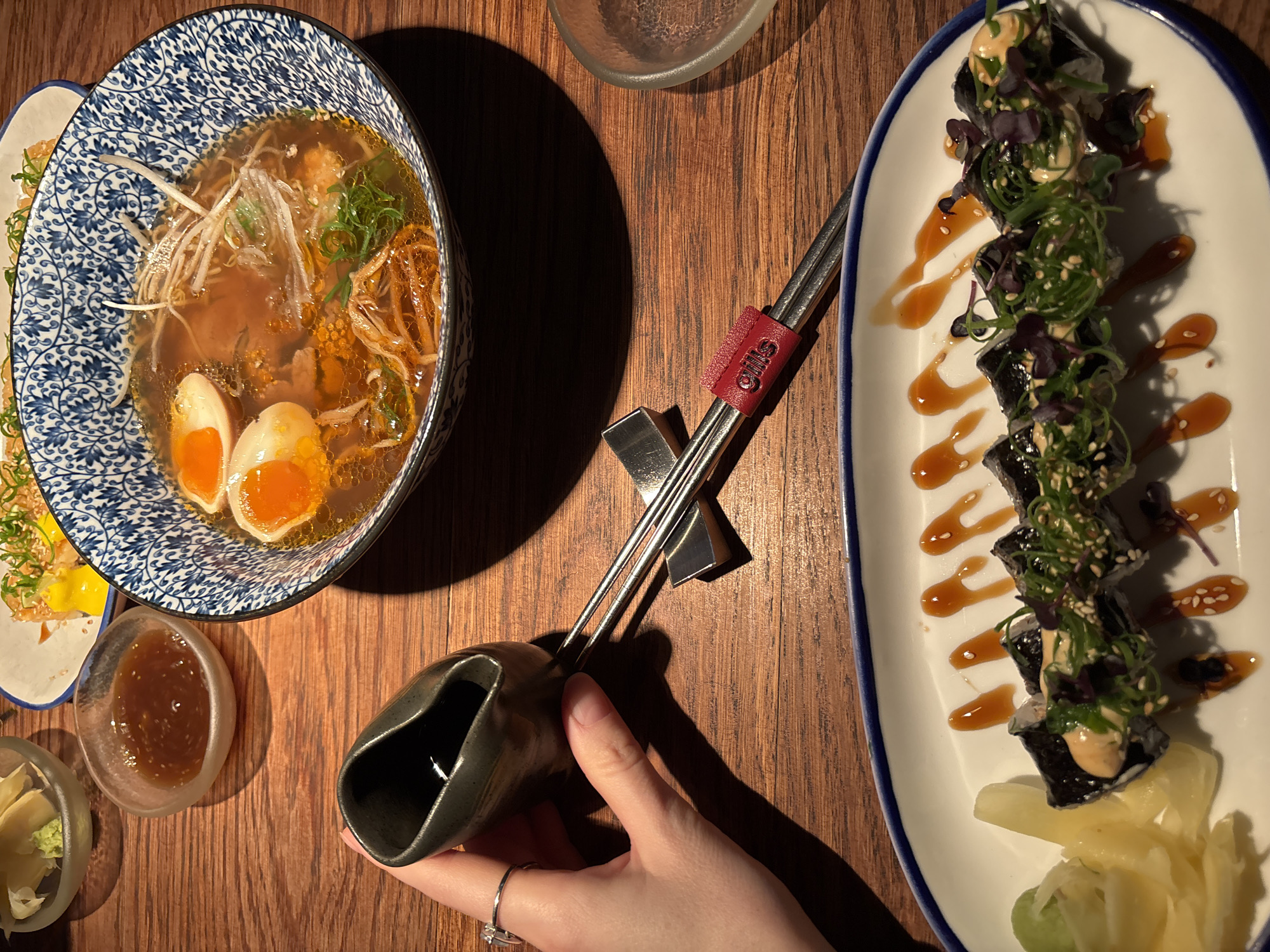 A top-down, indoor shot depicts a wooden table spread with food and utensils, likely in a restaurant. On the left, a blue-and-white patterned bowl contains ramen with a half-boiled egg. There is a small dish of dipping sauce and a small plate with ginger and wasabi. Silver chopsticks with a red chopstick rest are present. To the right, a white, oval plate holds a row of sushi rolls decorated with seaweed salad and sauce. A hand with a ring is holding a dark-colored sauce pouring vessel. The chopstick holder is from "Gills," a sushi chain. The presence of ramen and sushi suggests a Japanese restaurant. There is no indication it is in Saint Petersburg, Russia.