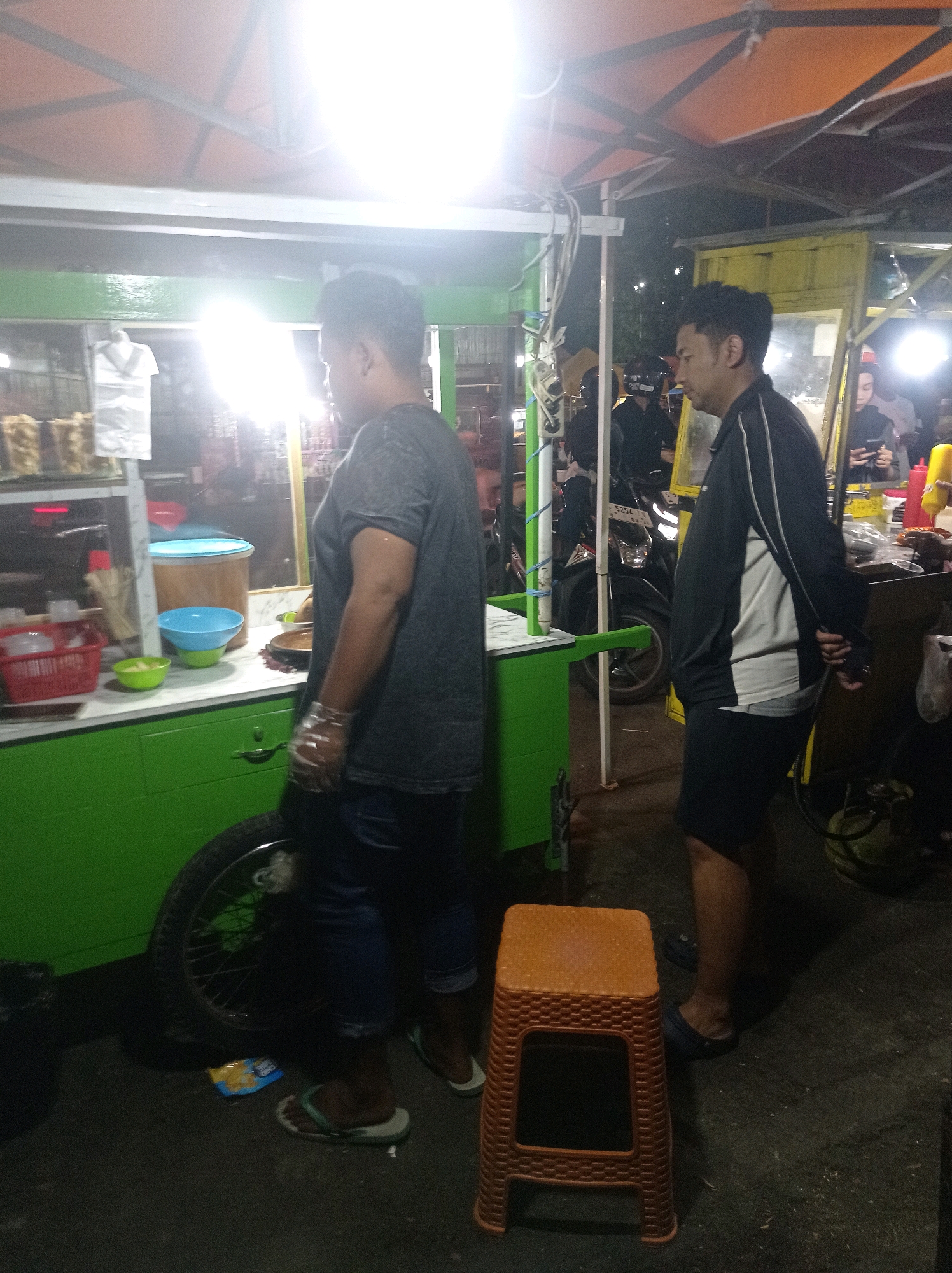 At a street food stall in Tuban, Indonesia, two men are visible in the foreground. The man on the left, facing away from the camera, is wearing a dark t-shirt and jeans and is wearing transparent plastic gloves. He is standing next to a green food cart. The man on the right, facing left, is wearing a black and white jacket and dark shorts. He is standing near a yellow food stall. Behind them, a motorcycle is parked and other people are visible in the background, suggesting a bustling evening scene. The food cart appears to be selling snacks or street food, with various containers and items displayed. The lighting suggests it is nighttime, with bright lights illuminating the stalls.