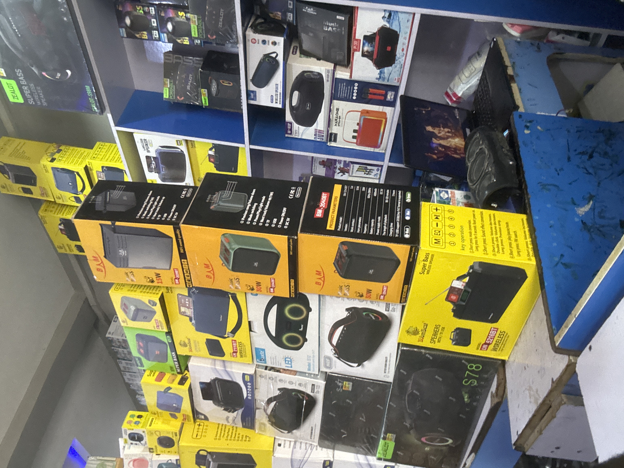 This media file is a close-up, high-angle shot of a retail display shelf filled with various electronic devices, primarily portable Bluetooth speakers and headphones. The items are predominantly boxed and stacked, with some individual units visible. The products are diverse in color and design, featuring brands like ZEALOT, BAM, and BoomBest. Many of the boxes display product names such as "SUPER BASS," "WIRELESS SPEAKER," and "KARAOKE SPEAKER," along with technical specifications like "15W," "30W," and "TWS." Some boxes also include graphics and feature lists highlighting functionalities like "LED," "Mode switching," and "Power ON/OFF."
The scene appears to be an electronics store or a market stall, likely in Jalingo, Nigeria, given the provided location context. The lighting suggests an indoor setting, possibly during daytime. There are no people visible in the frame. The focus is entirely on the merchandise, creating a sense of commercial inventory. The arrangement of the products suggests a retail environment where items are displayed for sale. Some of the boxes have Korean text, indicating a possible origin or manufacturing origin.