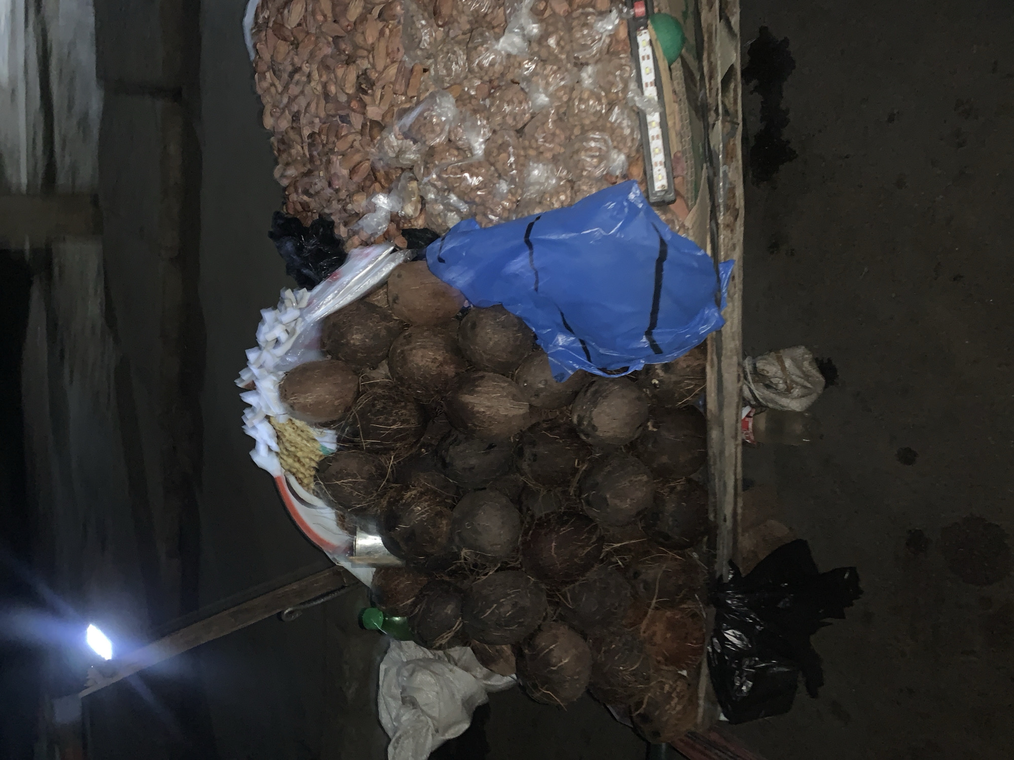 The image depicts a marketplace scene in Kabong, Nigeria, showcasing a vendor's wares. The primary objects visible are stacks of coconuts and bags of nuts. The coconuts are piled in a basket, partially covered by a blue plastic sheet. Adjacent to the coconuts, numerous bags of nuts are stacked. Additional plastic bags, some black, are scattered around the display. A measuring device is also present near the bags of nuts. The setting suggests an outdoor, potentially temporary, marketplace. The lighting is dim. No people are visible.