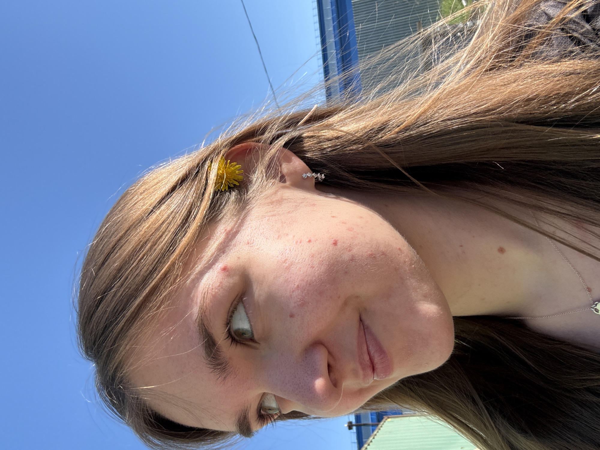 A young woman with light brown hair poses for a picture. A small, yellow flower adorns her ear. She wears a silver earring and a necklace. The clear, blue sky suggests a sunny day. A building is visible in the background. The photograph does not provide enough information to locate the specific location in Perm, Russia.