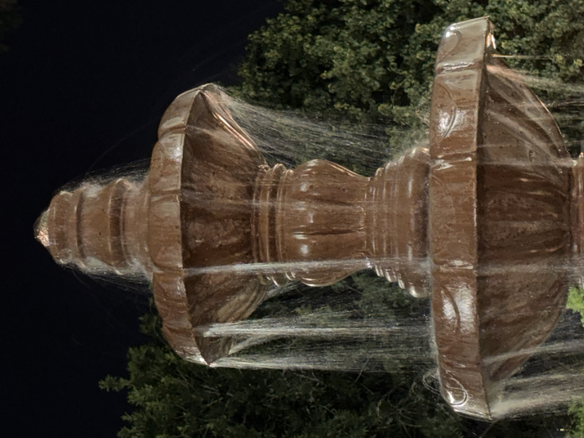 A close-up, low-angle, night-time shot features a multi-tiered, ornate, brown fountain with water cascading down its sides. The fountain is made of a stone-like material and has decorative carvings. Water streams down from the upper tiers, creating a misty effect. In the background, dark green foliage of trees is visible against a black night sky. The scene is located in Phoenix, United States of America.