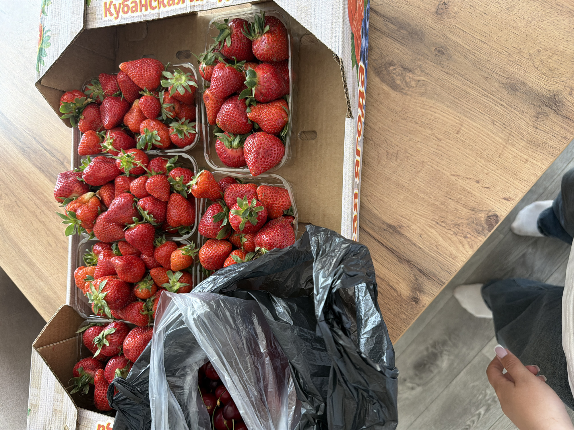 A cardboard box sits on a light brown wood-patterned table. Inside the box are several plastic containers filled with strawberries. A black plastic bag partially covers the strawberries and reveals some cherries underneath. To the right of the box is a person wearing grey jeans and white socks, holding their hand near the table's edge. The box is labeled "Кубанская." The scene appears to be indoors. The precise location within the city of Chaikovsky, Russia, is not discernible from the image.