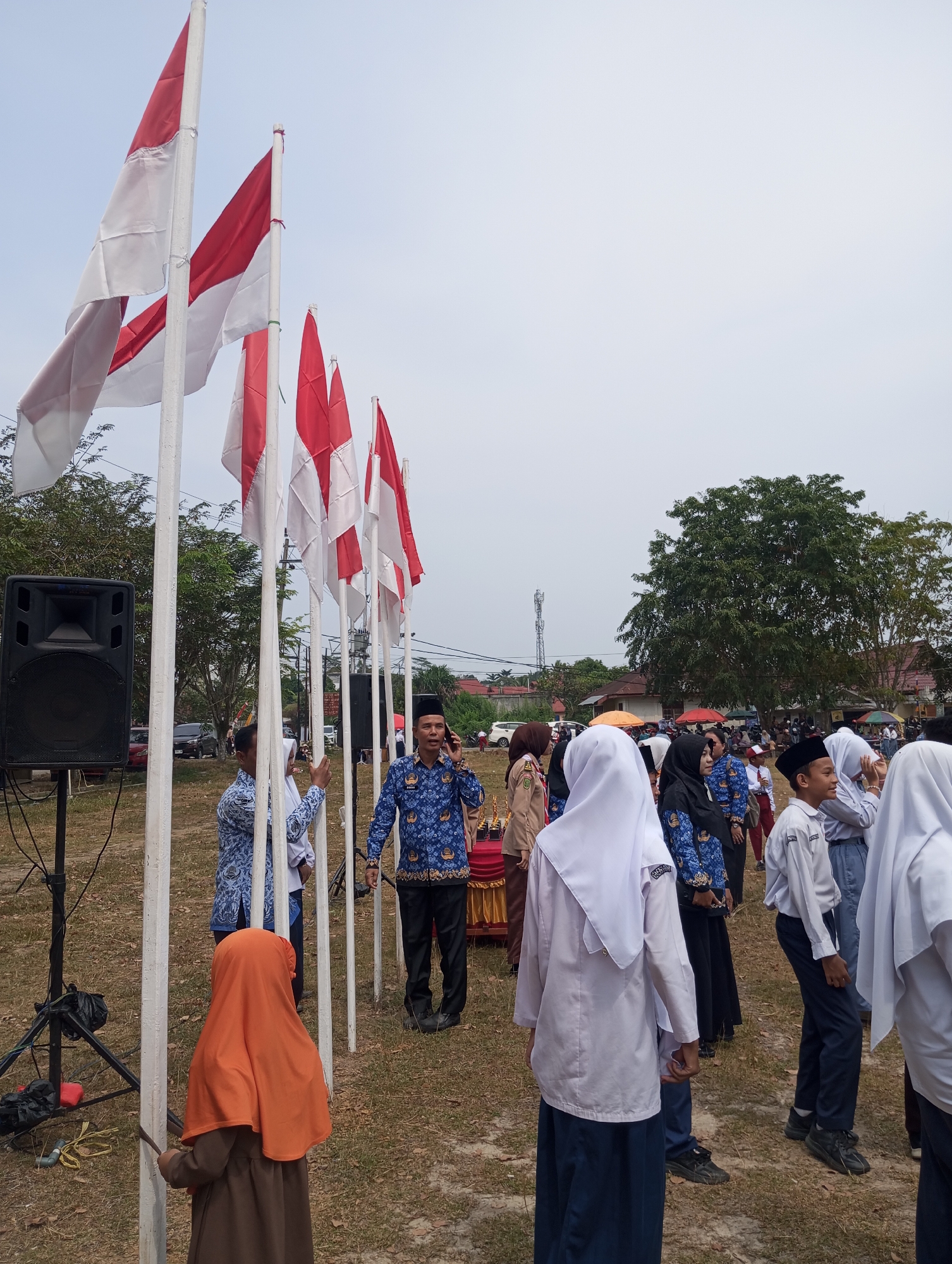 The image depicts an outdoor gathering, likely a school event, in the city of Pematang Reba, Indonesia. Several Indonesian flags are prominently displayed on white poles, waving in the breeze. A large black speaker is set up to the left of the flags.
In the foreground and midground, a diverse group of people are present. Many individuals are dressed in school uniforms, including white shirts and blue skirts or pants, and some wear headscarves. There is a man in a blue patterned shirt and black pants, speaking on a mobile phone. Another man in a similar blue patterned shirt stands near a table draped with a red cloth. A child in an orange headscarf and brown dress is also visible, standing near the flags.
The ground is covered with dry grass and some scattered leaves, indicating an open field or park. In the background, trees and buildings are visible, suggesting a semi-urban or rural setting within Pematang Reba. The sky is overcast and a communication tower can be seen in the distance. The overall scene suggests a formal event such as a flag-raising ceremony or a school assembly.