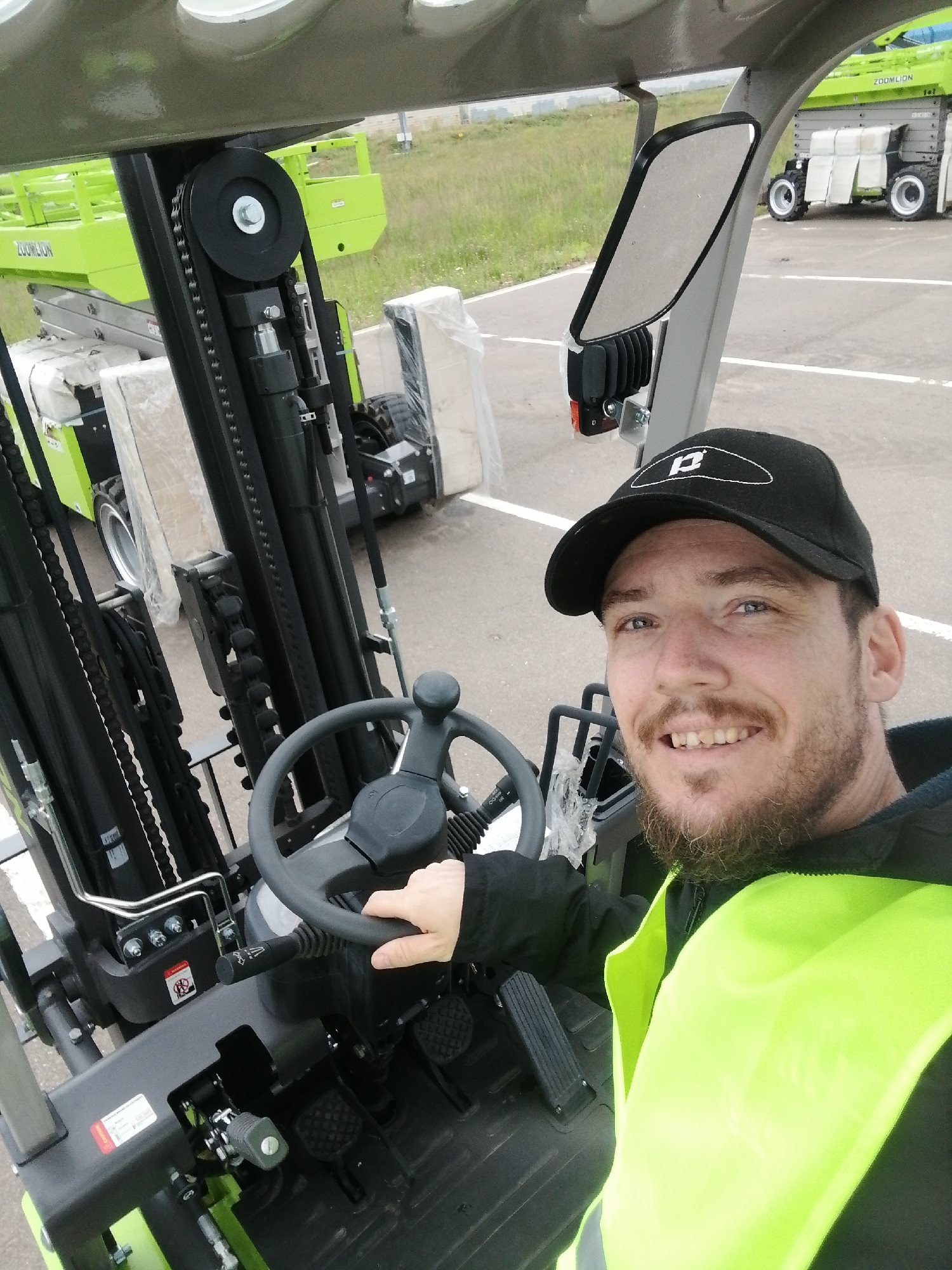 A man wearing a black baseball cap and a high-visibility yellow vest is sitting in the driver's seat of a forklift. He is smiling at the camera. The forklift's controls, including the steering wheel and pedals, are visible. In the background, another forklift is parked on a paved surface. The scene appears to be an outdoor industrial area, possibly a logistics yard or construction site, in the city of Spas-Kamenka, Russia.