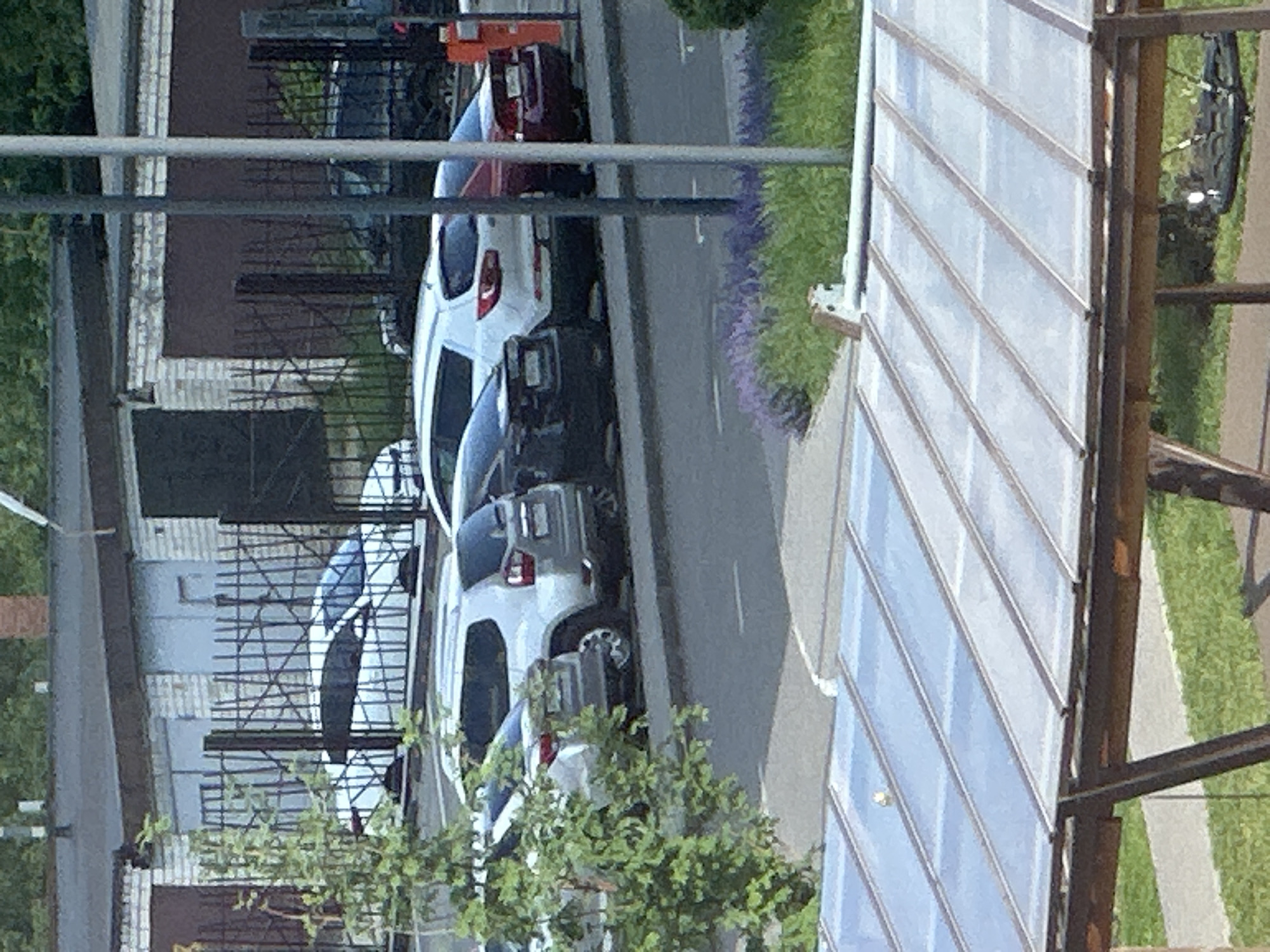 The image shows a parking lot with several cars. In the foreground, there is a transparent roof structure. In the background, there are garages and a fence. The cars visible include a white SUV, a silver SUV, a dark colored sedan, and a red sedan. The environment appears to be a residential area with trees and grass. There are no people visible in the image. The location is identified as Kazan, Russia.