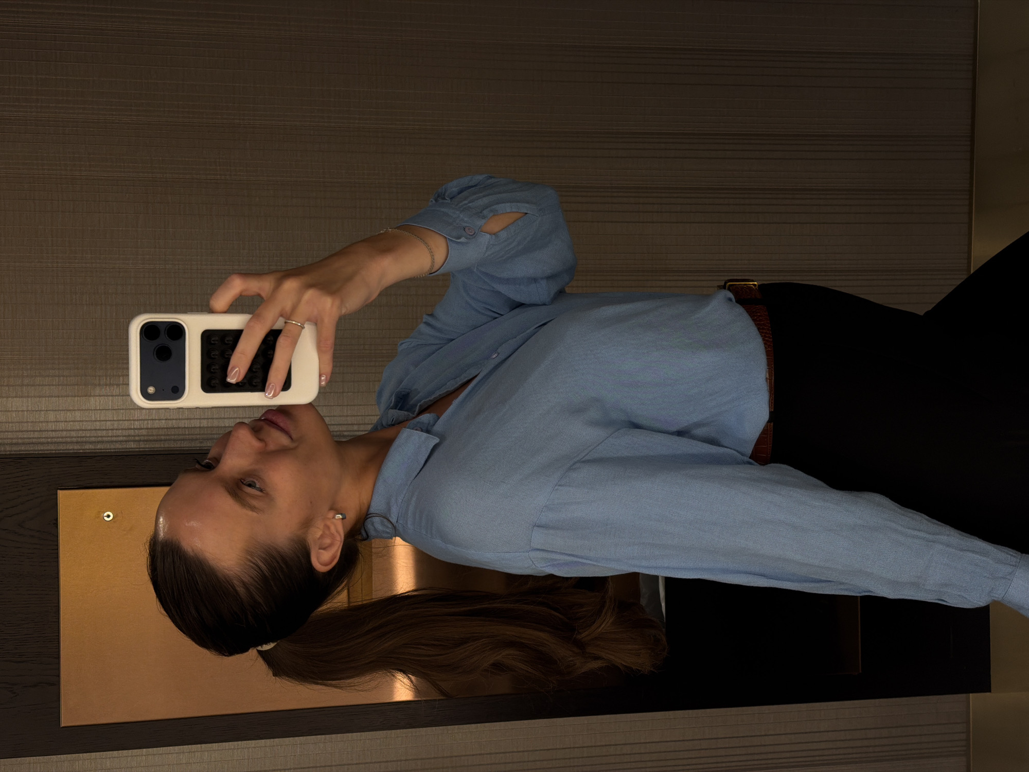 A woman is depicted taking a mirror selfie in an indoor setting. The image is rotated 90 degrees clockwise. She is holding a white smartphone with a dark camera module and what appears to be a black grip or accessory on the back. Her right hand, adorned with a simple ring on her ring finger and a thin bracelet on her wrist, holds the phone up, seemingly towards a reflective surface not fully visible but suggested by her reflection and posture.
The woman has dark hair pulled back into a ponytail, visible flowing over a dark, reflective surface that could be a table or counter. She is wearing a light blue long-sleeved button-up shirt with open cuffs, dark pants or a skirt, and a brown belt with a gold buckle. Her expression is calm, looking towards the phone's screen or her reflection. The background features a textured, horizontally-striped wall in a neutral dark brown tone, suggesting a modern interior space, possibly a hotel room or apartment. The lighting is subdued, typical of indoor artificial light. There is no discernible text visible in the image. The scene takes place in Budva, Montenegro.