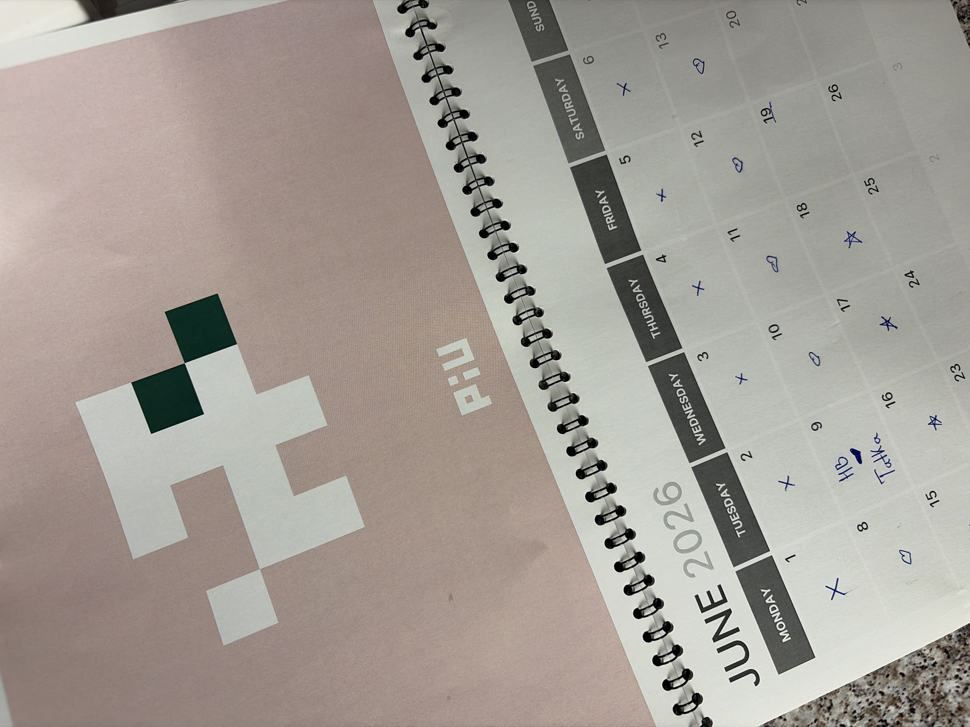 The image is a close-up of a calendar for June 2026. The calendar is spiral-bound and features a pink background with a pixelated white and green logo at the top. The days of the week are printed in gray boxes, with the current month and year, "JUNE 2026", displayed prominently. Some days have handwritten marks, including crosses, hearts, a star, and the text "HB Tatka" written on the 8th of June. The calendar is placed on a speckled countertop, likely a kitchen or dining area. The image does not contain any information about the city of Budva, Montenegro.