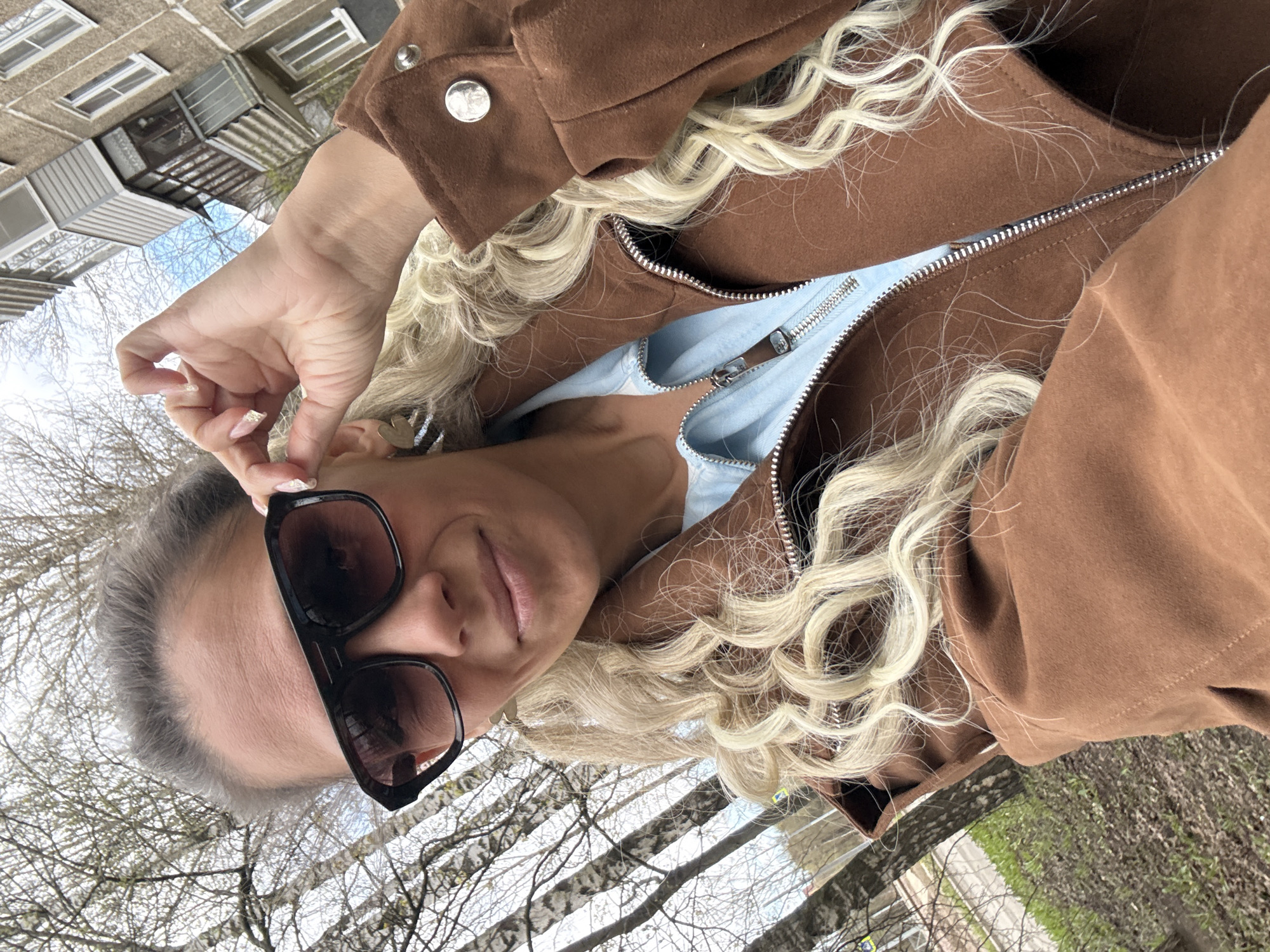 A person is captured in a selfie-style shot, slightly smiling and wearing large, black sunglasses that obscure their eyes. They have long, predominantly blonde and curly hair, with some gray hair visible near their temples. Their hand, adorned with long, light-colored nails possibly featuring glitter or decoration, is raised near their ear. Small, possibly heart-shaped earrings are visible on their ears.
The individual is dressed in a brown, suede-like jacket with a silver zipper and silver snap buttons on the lapel. Underneath, a light blue top with a zipper is visible.
The setting is outdoors, suggesting a street or park area, characteristic of Rusanovo, Russia, as per the provided context. In the background, a multi-story, possibly concrete apartment building with multiple windows and balconies is visible. The foreground and midground feature numerous deciduous trees with bare branches, indicating late winter or early spring. Patches of green grass are visible on the ground. The sky appears overcast or partly cloudy, suggesting daytime. No visible text is present in the image.