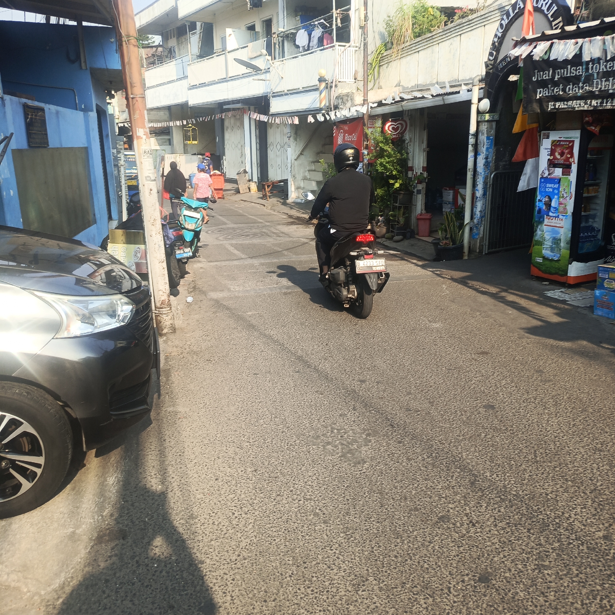 The image depicts a street scene in South Jakarta, Indonesia, likely a residential or mixed-use area with shops. In the foreground, the front of a dark-colored car is visible on the left. A person wearing a black helmet and black clothing is riding a motorcycle down the street, casting a shadow on the asphalt. Other people and motorcycles are visible further down the street, and buildings line both sides of the road. Some of the buildings appear to be shops with signage and displays. The sky is bright, suggesting it is daytime.