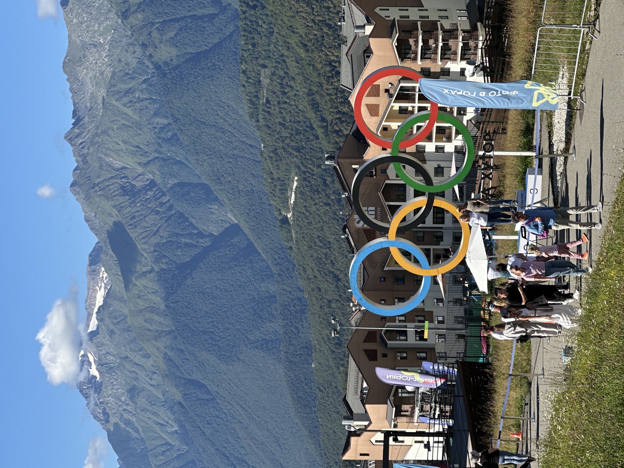The image captures a sunny day at a resort in Estosadok, Russia, with majestic mountains as a backdrop. In the foreground, a group of people are gathered near a large Olympic rings monument. The rings are colorful, with blue, yellow, black, green, and red rings prominently displayed. Behind the monument, several multi-story buildings with brown and beige facades are visible, suggesting a resort or residential complex. A blue flag with text and a yellow symbol stands to the right of the Olympic rings. People are walking and standing around, some are posing for photos. The ground is a paved path with grassy areas on either side. In the background, the imposing mountains are covered with lush green trees, and their peaks are capped with snow. The sky is clear blue with a few fluffy white clouds.