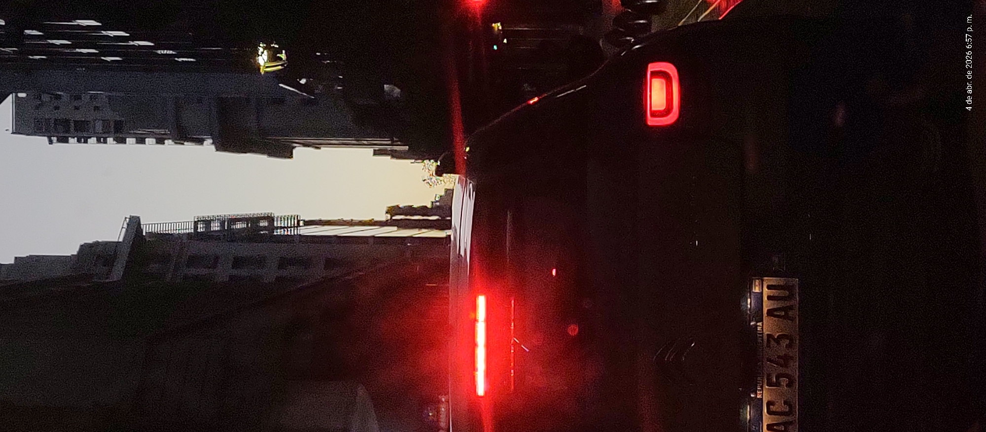 The image shows a dark urban scene in Buenos Aires, Argentina at 6:57 p.m. on April 4, 2026. Buildings are visible in the background. A black car with a lit "D" tail light and vertical red light is in the foreground. The car's license plate reads "AC 543 AU". The overall lighting is poor.