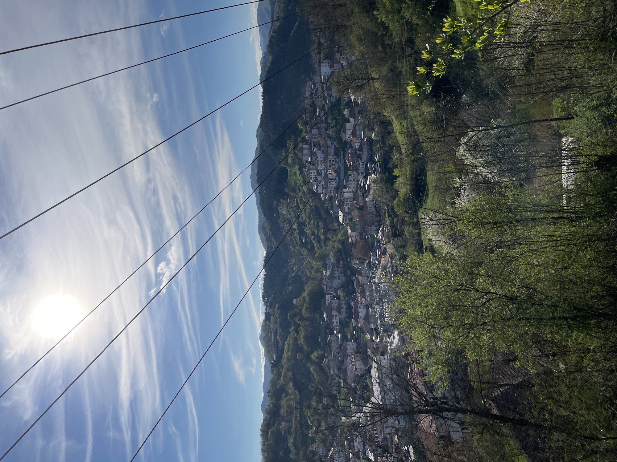 This image captures a vibrant, high-angle landscape view of Zlatograd, Bulgaria, showcasing a town nestled in a verdant mountainous valley under a clear sky. The perspective is from an elevated vantage point, looking down and across the scene.
In the upper left quadrant, the sun shines brightly, creating a prominent glare and illuminating the scene with strong daylight, suggesting either morning or late afternoon. The sky is a brilliant blue, adorned with delicate, wispy white clouds that stretch across the expanse. Several dark, parallel lines, likely utility or power cables, traverse the upper half of the frame diagonally, cutting across the sky and extending over the valley.
The immediate foreground features a dense scattering of trees and foliage. Many trees display fresh, bright green leaves, indicating the spring season, while some deciduous trees still have bare branches, adding textural variety.
Below the foreground foliage, the main subject is the picturesque town of Zlatograd. It comprises numerous white-washed buildings, each topped with characteristic orange or reddish-brown tiled roofs. The structures are tightly clustered and appear to follow the natural contours of the valley, extending up the lower slopes of the surrounding hills.
Beyond the town, the landscape transitions into a series of undulating hills and mountains, which are densely forested with a rich variety of green trees. The distant mountain ridges are softly visible on the horizon, hinting at the vastness of the natural environment.
No explicit human activity is visible within the frame, and there is no discernible text. The image conveys a sense of tranquility and natural beauty, characteristic of a serene mountain community.