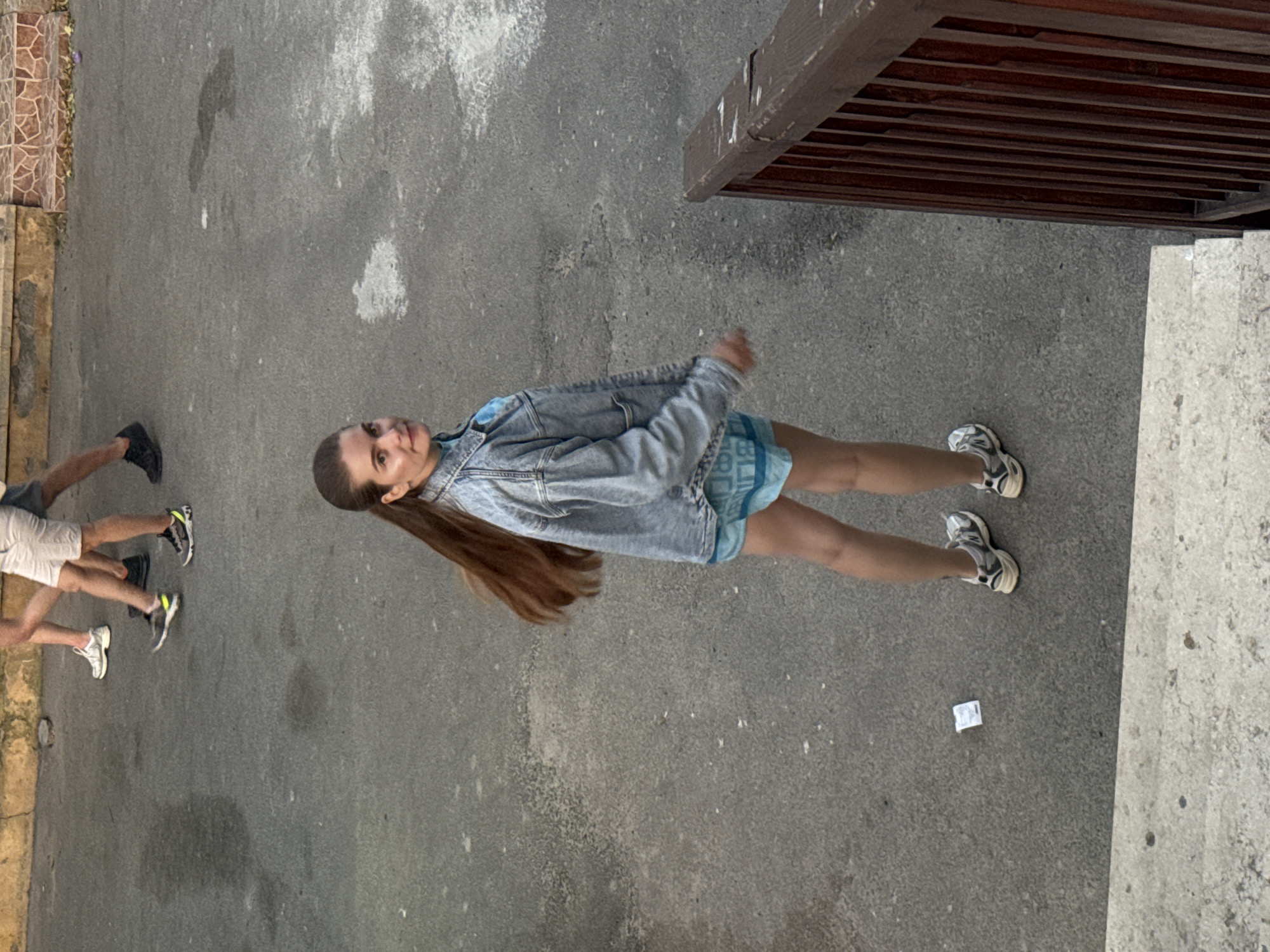 A young woman stands on an asphalt surface, her body oriented vertically in the image. She is wearing a light blue denim jacket over a blue top and light blue shorts. Her long, brown hair is pulled back into a ponytail. She is wearing white and grey athletic shoes. Her right arm is bent at the elbow, and her left arm is extended slightly to her side. She is looking towards the camera.
To the left of the woman, two other people's legs are visible. The person closer to the woman is wearing white shorts and white athletic shoes. The person further left is wearing dark pants or shorts and black shoes.
The setting appears to be outdoors, possibly a street or walkway, with an asphalt ground. To the right of the woman, a wooden structure with horizontal slats is visible, possibly part of a deck or balcony. A concrete curb or step is also present on the far right of the image. The background shows some textured tiling on the left and the continuation of the asphalt surface.
There is a small, white piece of paper on the asphalt near the woman's feet. The lighting suggests it is daytime, but it's difficult to determine the exact time or weather conditions. No text is clearly visible on any of the subjects or objects. The overall scene has a casual, everyday feel.