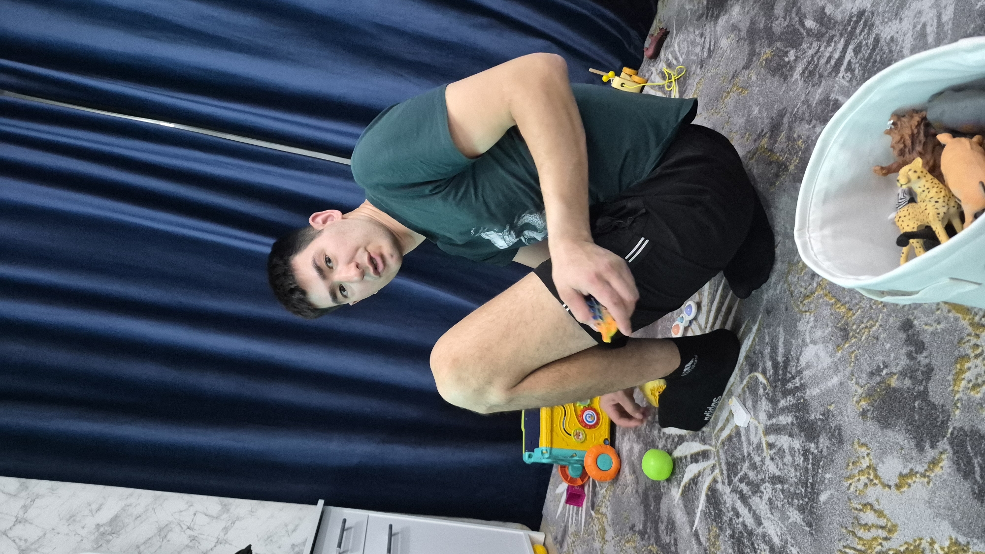 The scene is an interior view of a room, likely a residence, located in Chaikovsky, Russia. An adult male is crouched on a grey patterned rug with abstract white and yellow/gold designs. He has short dark hair and is wearing a dark green t-shirt with a graphic, black athletic shorts with two white stripes on the left leg, and black socks with "adidas" branding visible on the left foot. He holds a small orange and blue toy in his right hand and looks directly at the camera. A bright green spherical toy is on the rug to his left. Behind him, a yellow and blue multi-sided activity cube with orange wheels is present. In the bottom right foreground, a light blue fabric bucket contains several plastic animal figures, including a leopard, zebra, and lion. The background consists of a full-height dark blue curtain, and to the far left, a white dresser or cabinet with two silver handles is visible against a wall with a white and grey marble-like pattern.
