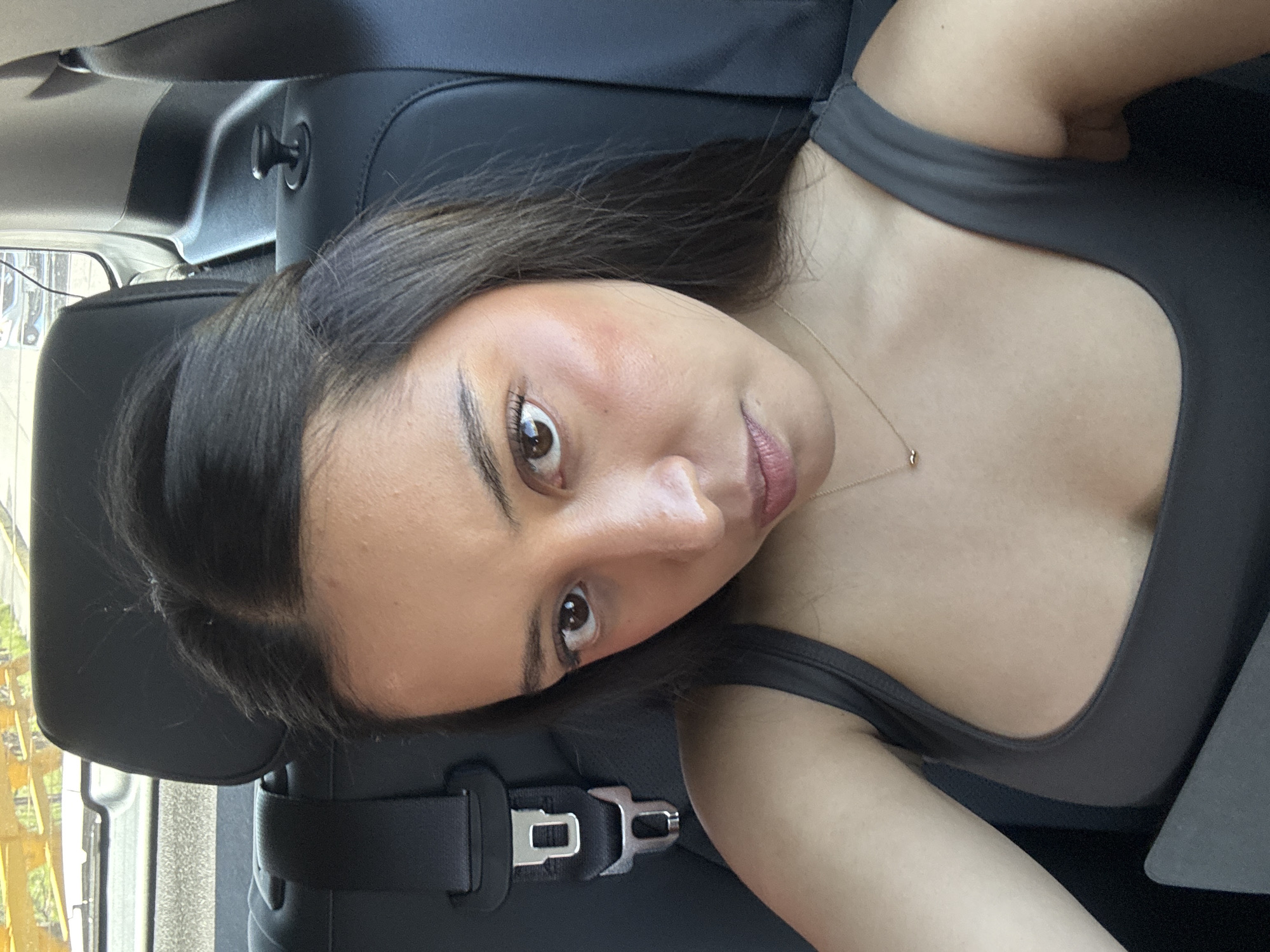 A portrait-style selfie captures a young woman inside a car, likely in the back seat, in San Bernardino Tlaxcalancingo, Mexico. The image is taken from a slightly low angle, with the woman's head tilted as she looks directly at the camera.
The subject is a young woman with long, dark brown hair that falls over her shoulders. She has dark eyes, well-defined eyebrows, and appears to be wearing subtle makeup, including a pinkish blush on her cheeks and a natural lip tint. She is dressed in a dark grey or charcoal-colored tank top or camisole, and a delicate gold chain necklace with a small, minimalist pendant adorns her neck.
The setting is the interior of a car, characterized by black leather seats. A prominent black seatbelt buckle is visible to her left, partially obscured by her hair, suggesting she is seated and possibly belted. A black coat hook is also visible on the car's interior trim above and behind her head. Through the blurry car windows in the background, natural daylight is evident, indicating the photo was taken during the day. The out-of-focus exterior shows hints of light-colored structures or buildings and some green foliage. The woman's expression is calm and composed. There is no visible text in the image.