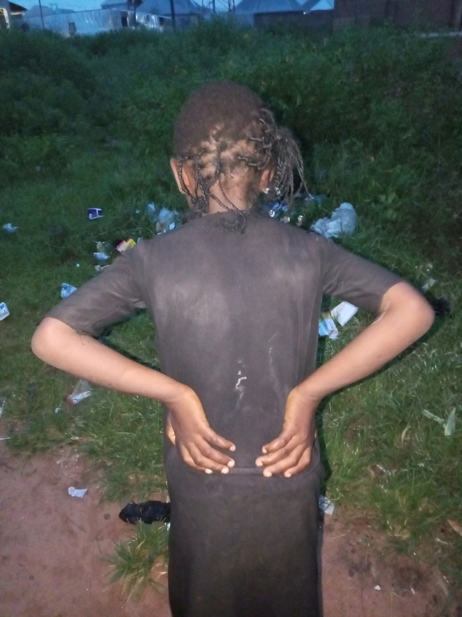 A person stands with their back to the camera in a grassy, outdoor area littered with trash. The person is wearing a dark t-shirt and their hair is styled in braids. Their hands are placed on their lower back. The background shows a slightly overcast sky and some buildings in the distance, suggesting an urban or semi-urban environment. The lighting indicates it is either early morning or late evening. The presence of scattered waste suggests a disregard for the environment. The overall scene appears to be in Tempure, Nigeria. No text is visible in the image.