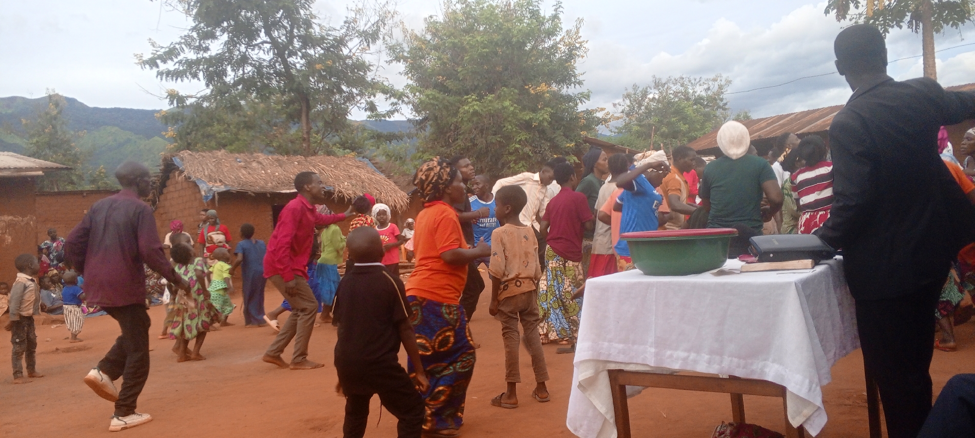 A group of people is gathered in an open dirt area in Ngalula, Democratic Republic of the Congo. Behind them are trees, and mountains in the distance are partially obscured by clouds. To the left, there are several small structures made of mud bricks, with straw roofs. People are milling about, some dancing, and others are standing and observing. In the foreground, there is a white table with a green bowl, a black book, and a light-colored book on top of it. A person in a black suit stands partially in front of the table with their back facing the camera. The people in the crowd wear a variety of colored shirts and dresses.