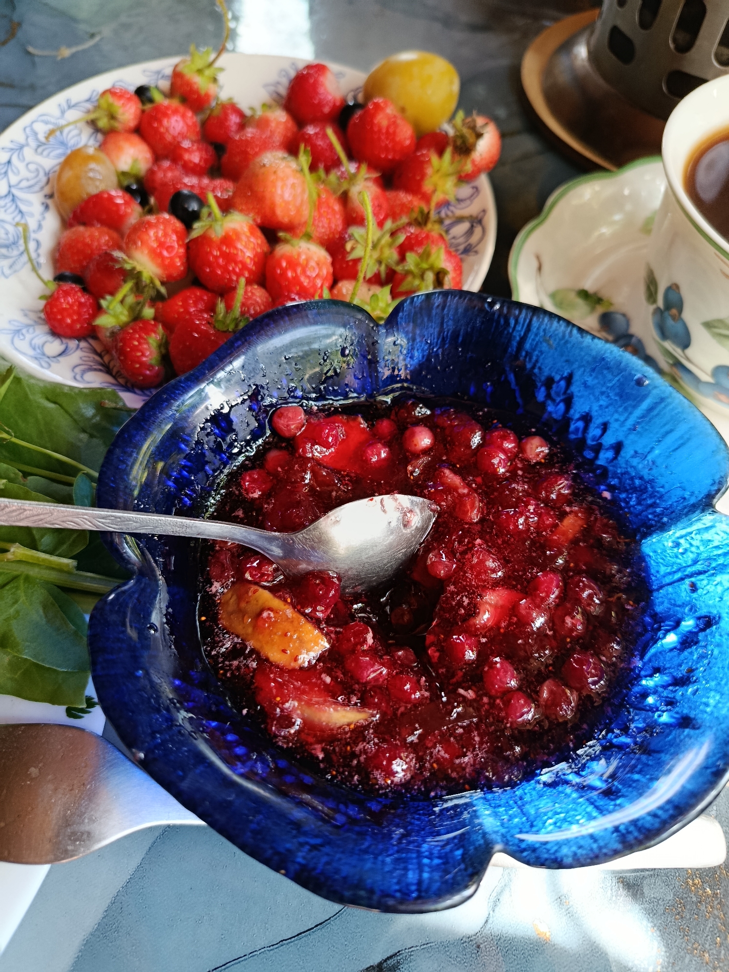 An overhead shot shows a close-up of a vibrant, blue, flower-shaped bowl filled with a glistening red berry compote. A silver spoon is partially submerged in the compote. To the left of the blue bowl are large green leaves, possibly spinach or watercress. Behind the compote bowl, to the left, is a white plate filled with fresh strawberries and a few blueberries. A lemon is visible on the plate among the berries. To the right of the compote bowl is a white teacup with a blue floral pattern, filled with dark coffee. The teacup sits on a saucer with the same blue floral pattern. A dark metal object, possibly a French press or coffee grinder, is visible in the background behind the teacup. The scene is set on a textured, pale blue and gray surface, possibly a table. The lighting suggests an outdoor setting, with bright daylight casting subtle shadows. The location is Unknown, Unknown.