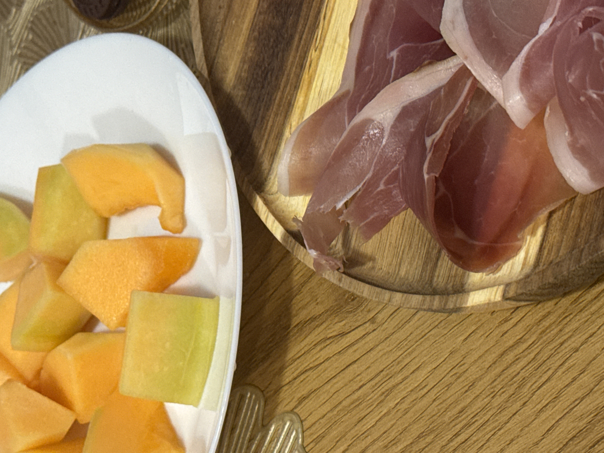 A close-up, overhead shot shows a white plate of sliced cantaloupe and honeydew melon next to a wooden board with slices of prosciutto. The fruit is cut into triangular wedges. The prosciutto slices are thin and layered. The background is a wooden surface with visible grain. The scene appears to be indoors, possibly in a kitchen or dining area.