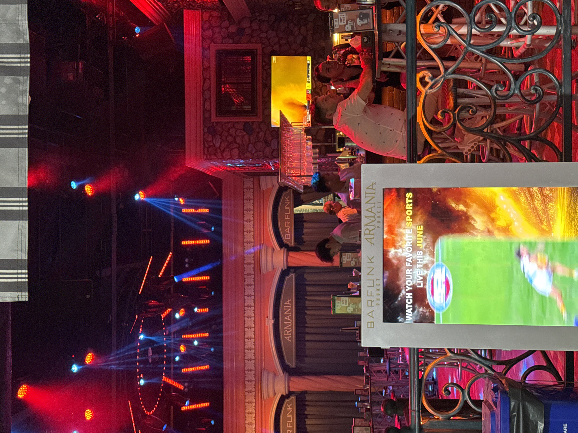 This image captures a lively scene inside Bar Funk in Patong, Thailand, likely during an evening event. Red and blue stage lights illuminate the area, creating a vibrant atmosphere. In the foreground, a large sign displays "WATCH YOUR FAVORITE SPORTS LIVE THIS JUNE" with a graphic of a soccer game. The bar area is visible, with patrons seated at high tables and stools, some engaging in conversation. Behind the bar, a wall features stone-like textures and a television screen showing a sports broadcast. The overall environment suggests a bustling bar or club setting with a focus on entertainment and sports viewing.