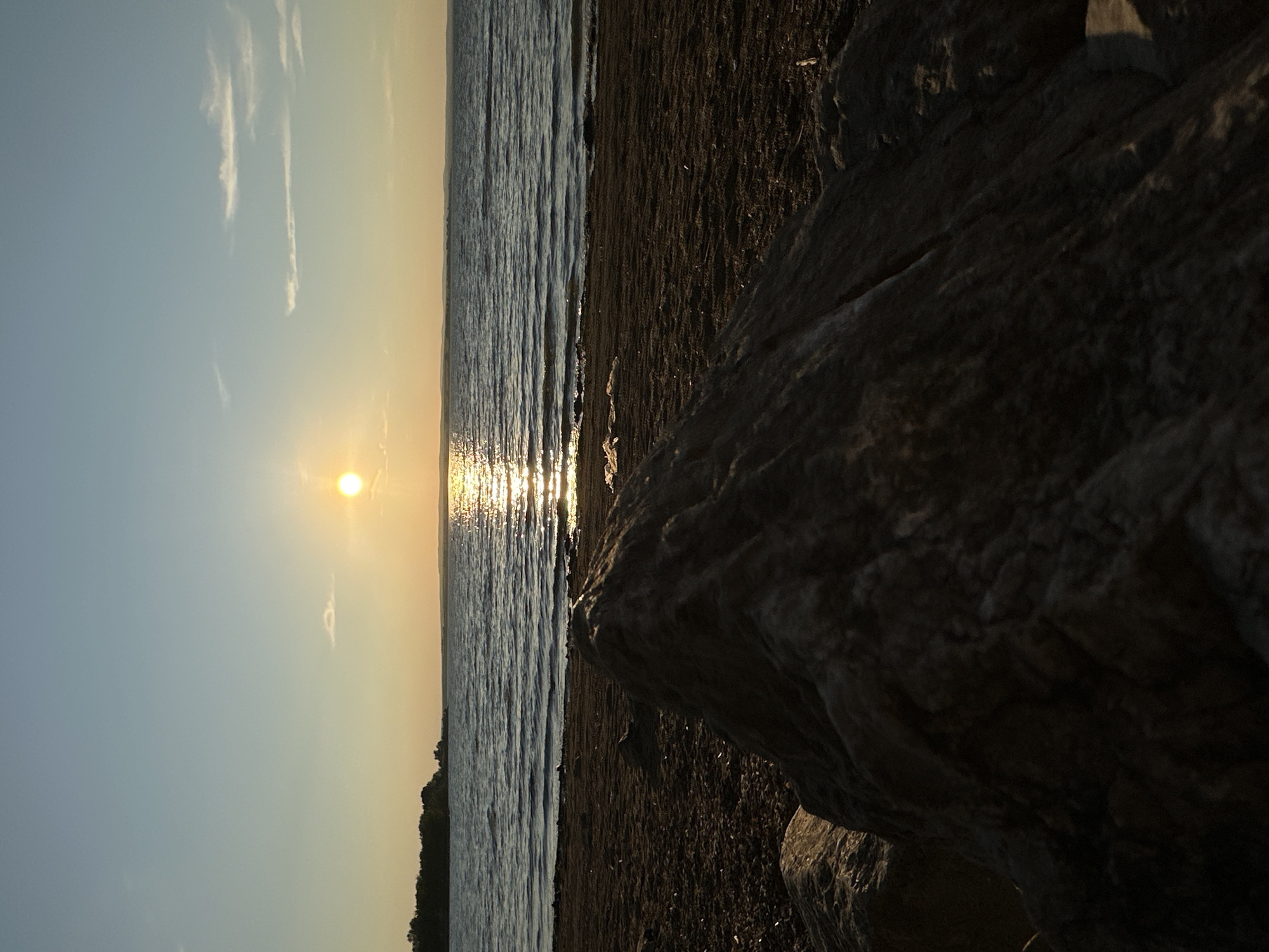 The image depicts a sunset over a body of water, likely a reservoir, near Energetik, Russia. In the foreground, large rocks and sand create a beach-like setting. The sun is low on the horizon, casting a bright reflection across the rippling water. In the distance, a treeline is visible, suggesting a wooded area on the opposite shore. The sky is a gradient of pale blue and warm yellow, with a few wispy clouds. The overall scene is serene and natural, characteristic of a lakeside environment at dusk. There are no people or animals visible in the image.