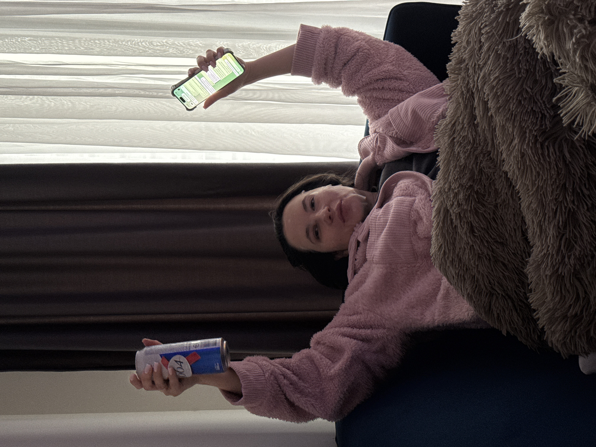 A woman reclines on a dark blue couch, covered partially by a large, brown, furry blanket. She is wearing a pink, fuzzy sweater, and holding a can of Red Bull in her right hand and a smartphone, displaying a messaging app, in her left. A white, horizontal-slat blind is visible behind her, with a brown curtain below. The location is likely indoors, within a building in Belgrade, Serbia, based on the can of Red Bull, which is available for purchase there.