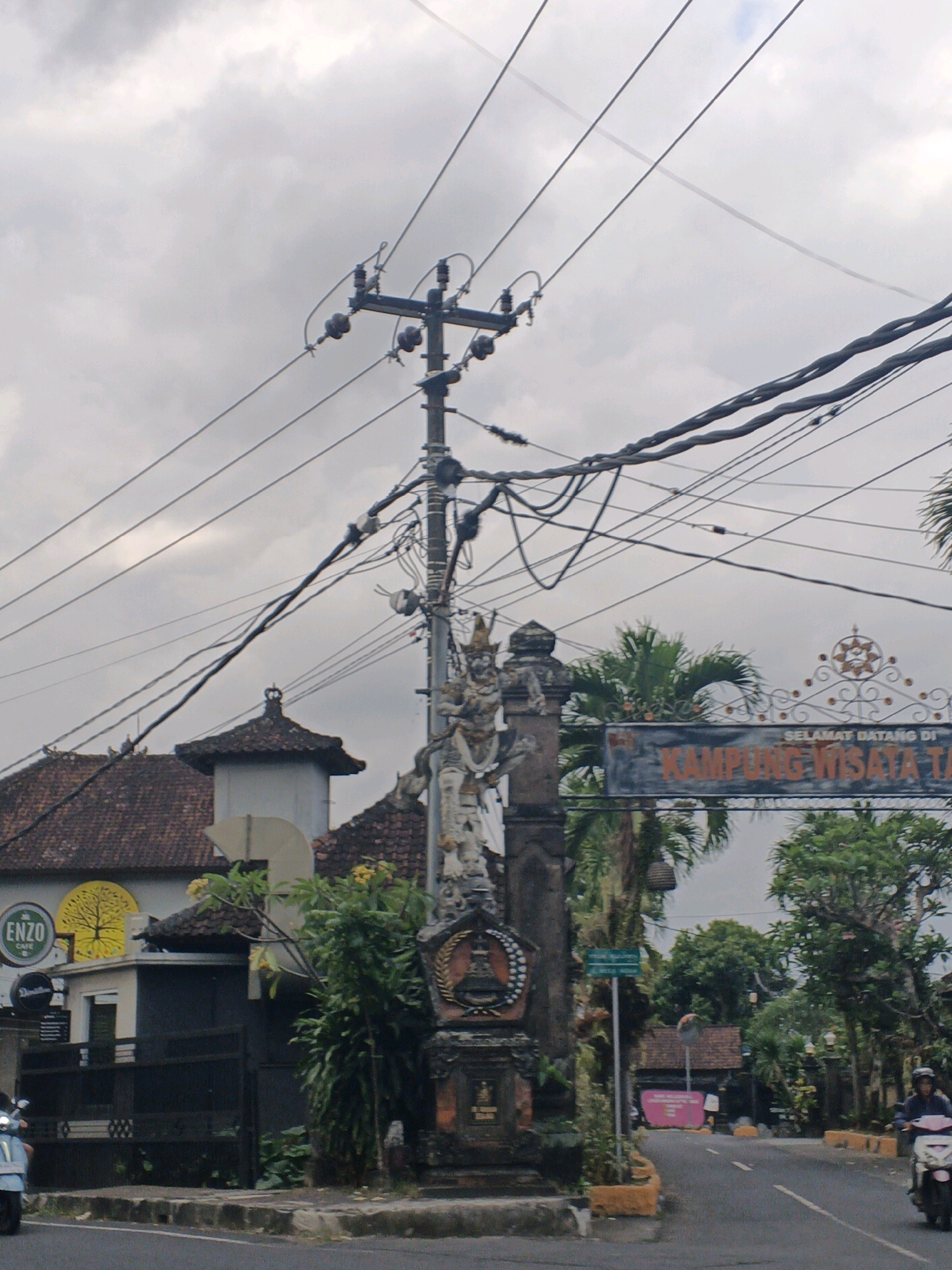 The image depicts a street scene in Ubud District, Indonesia, under an overcast sky. A prominent feature is a tall utility pole with a complex arrangement of electrical wires, creating a dense network overhead. To the right of the pole, an ornate gate with decorative metalwork stands, bearing text that reads "SELAMAT DATANG DI KAMPUNG WISATA TA," translating to "Welcome to Tourist Village." Adjacent to the gate is a stone monument or statue, featuring intricate carvings and what appears to be a traditional Balinese architectural element at its base.
On the left side of the street, a building with a tiled roof and a lighter-colored facade is visible. A sign for "ENZO CAFE" with a yellow circular logo depicting a tree is affixed to this building. A dark fence runs along the front of the property.
The road itself is paved, with a gray asphalt surface. A light blue scooter is partially visible on the left side of the road, positioned behind the fence. On the right side of the road, a person is riding a white scooter, moving away from the viewer. Lush green trees and foliage are abundant throughout the scene, contributing to the tropical atmosphere. The overall impression is one of a bustling yet traditional street in a tourist-oriented area.